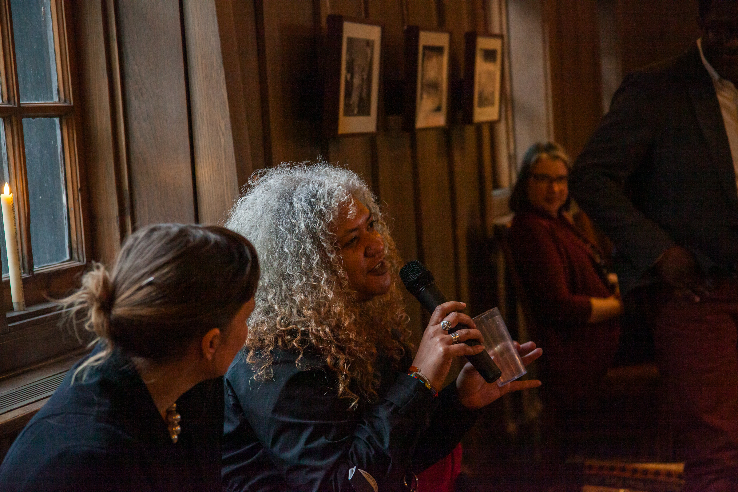 A woman with curly gray hair speaks into a microphone while seated indoors, next to another woman. Framed photos hang on the wooden-paneled wall, and two other people sit in the background. Warm lighting fills the room.