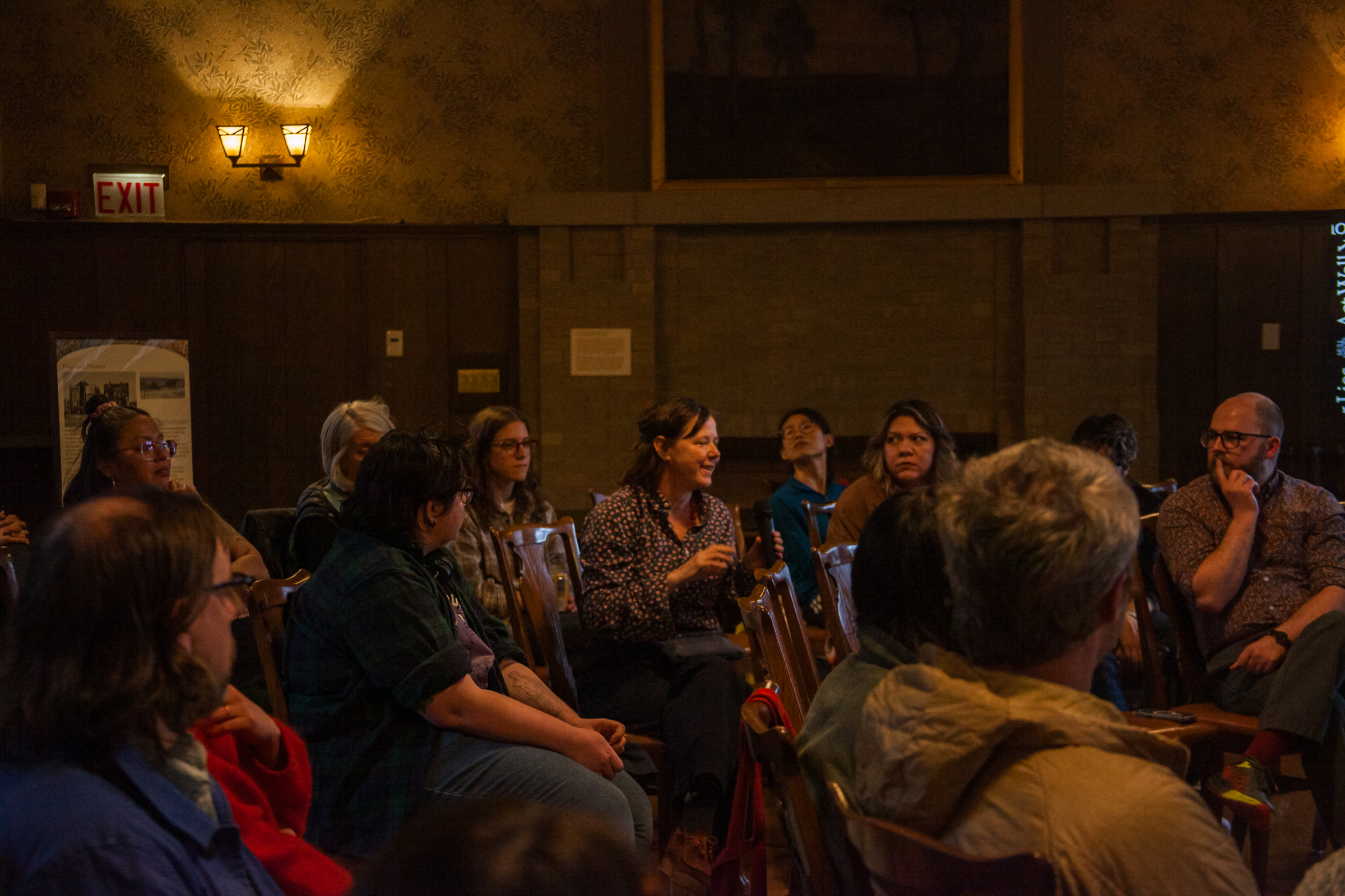 A group of people sit in a dimly lit room, attentively listening to a woman who is speaking and gesturing with her hands. The atmosphere appears to be serious and focused, suggesting a discussion or meeting.