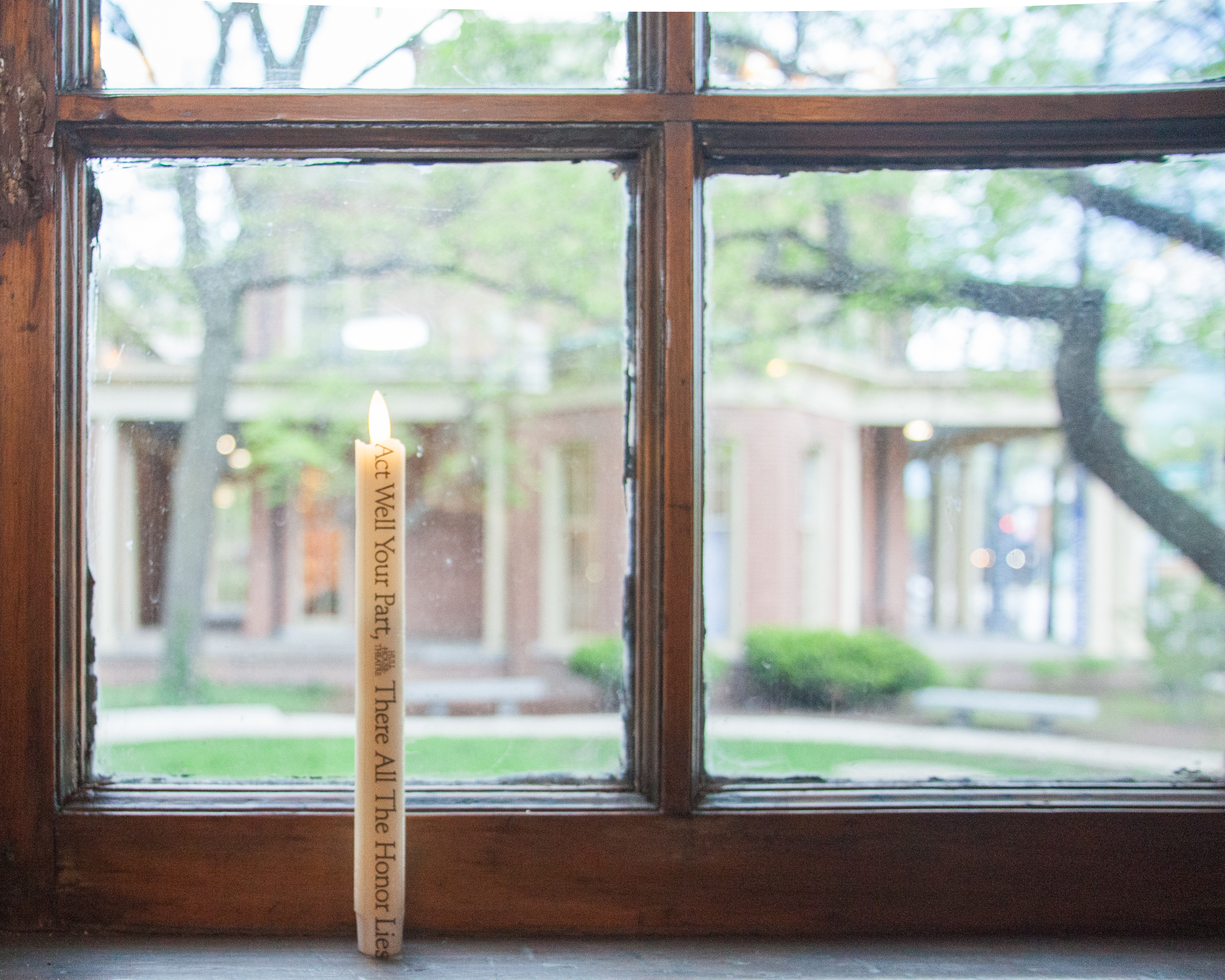 A lit candle with text on it sits on a windowsill in front of a wooden-framed window, looking out onto a courtyard with trees and brick buildings in the background.