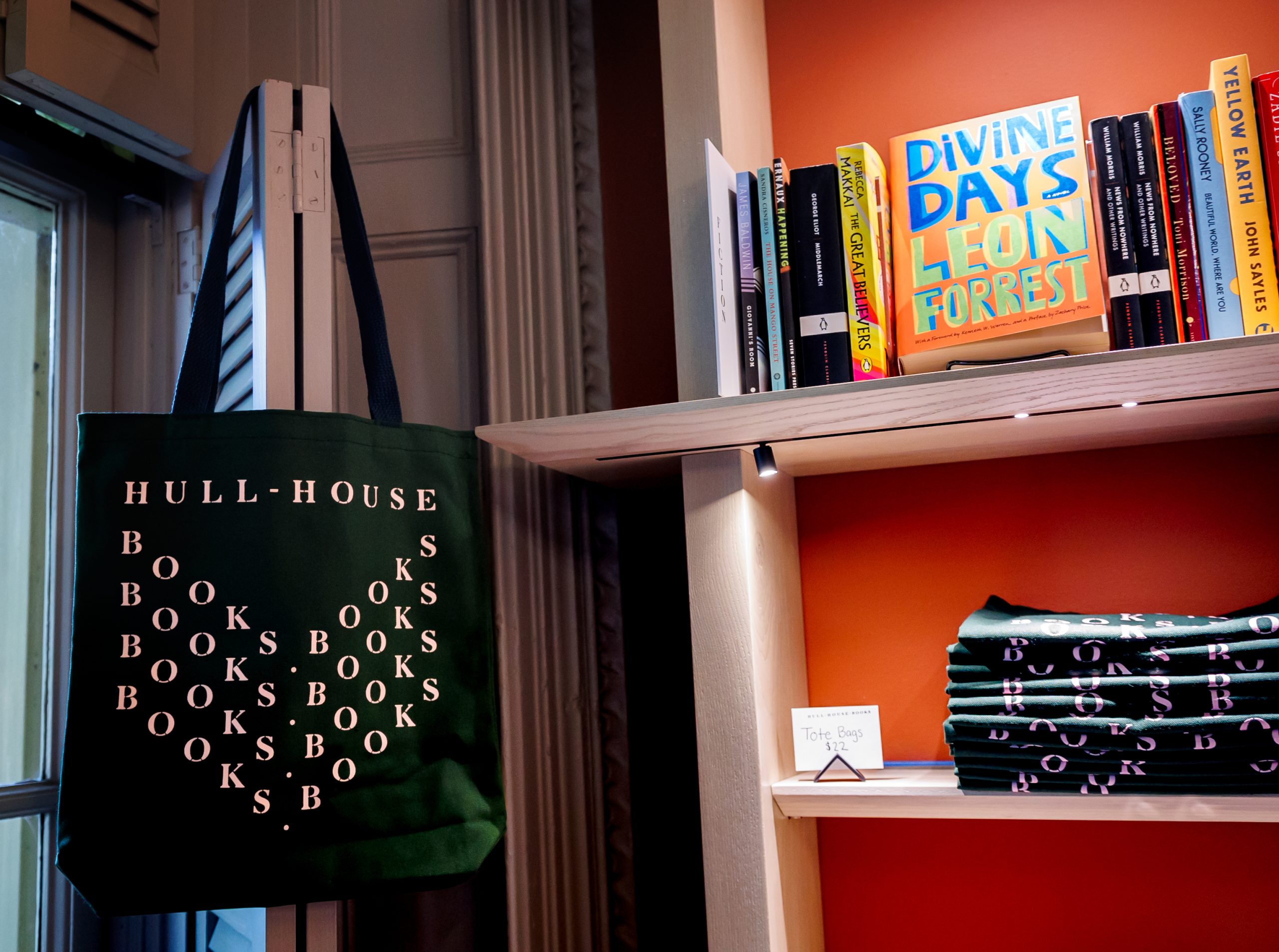 A dark green tote bag reading HULL-HOUSE BOOKS hangs by a window beside orange bookshelves displaying folded totes, books, and a featured book titled DIVINE DAYS by Leon Forrest.