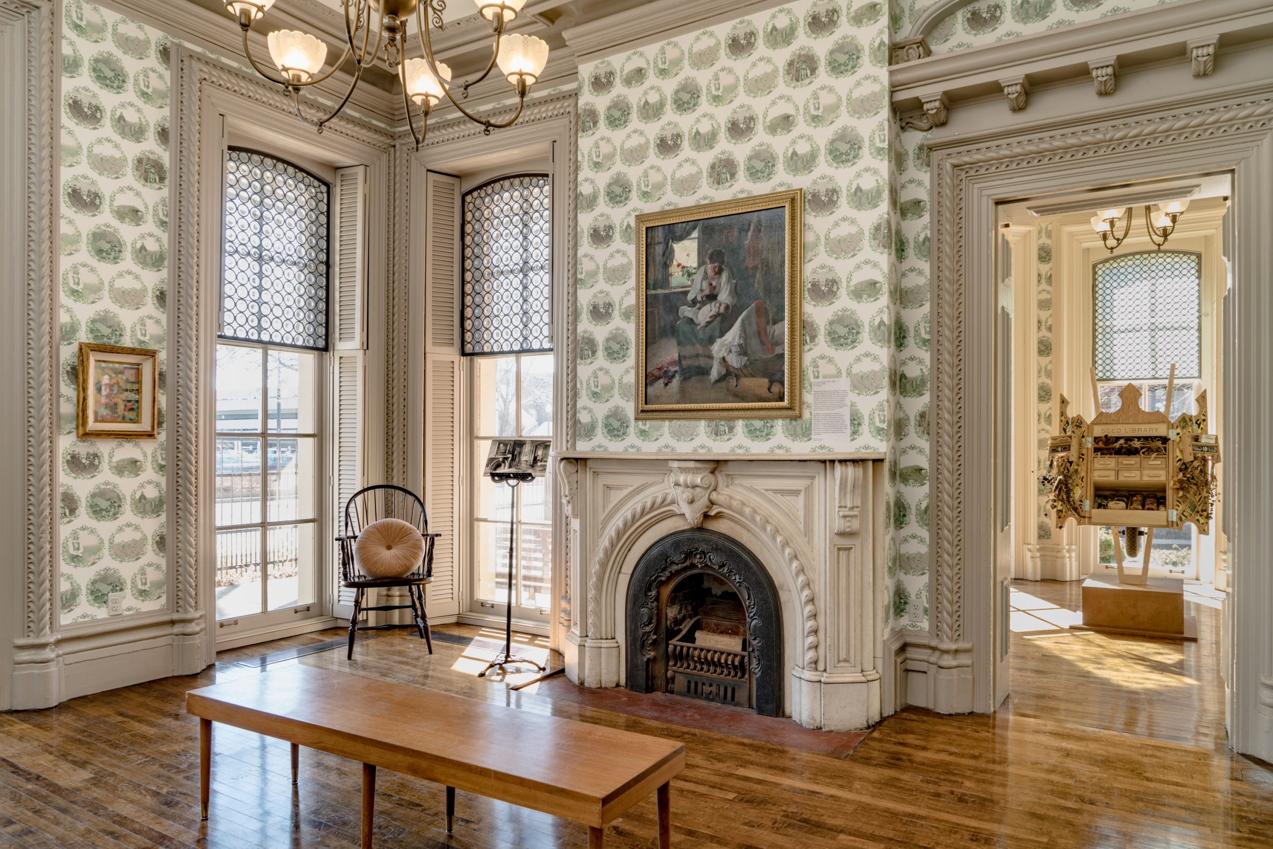 A historic room with ornate wallpaper, large windows, a decorative fireplace, wooden floors, a wooden bench, and paintings on the walls. An open doorway reveals another decorated room with an elaborate cabinet.