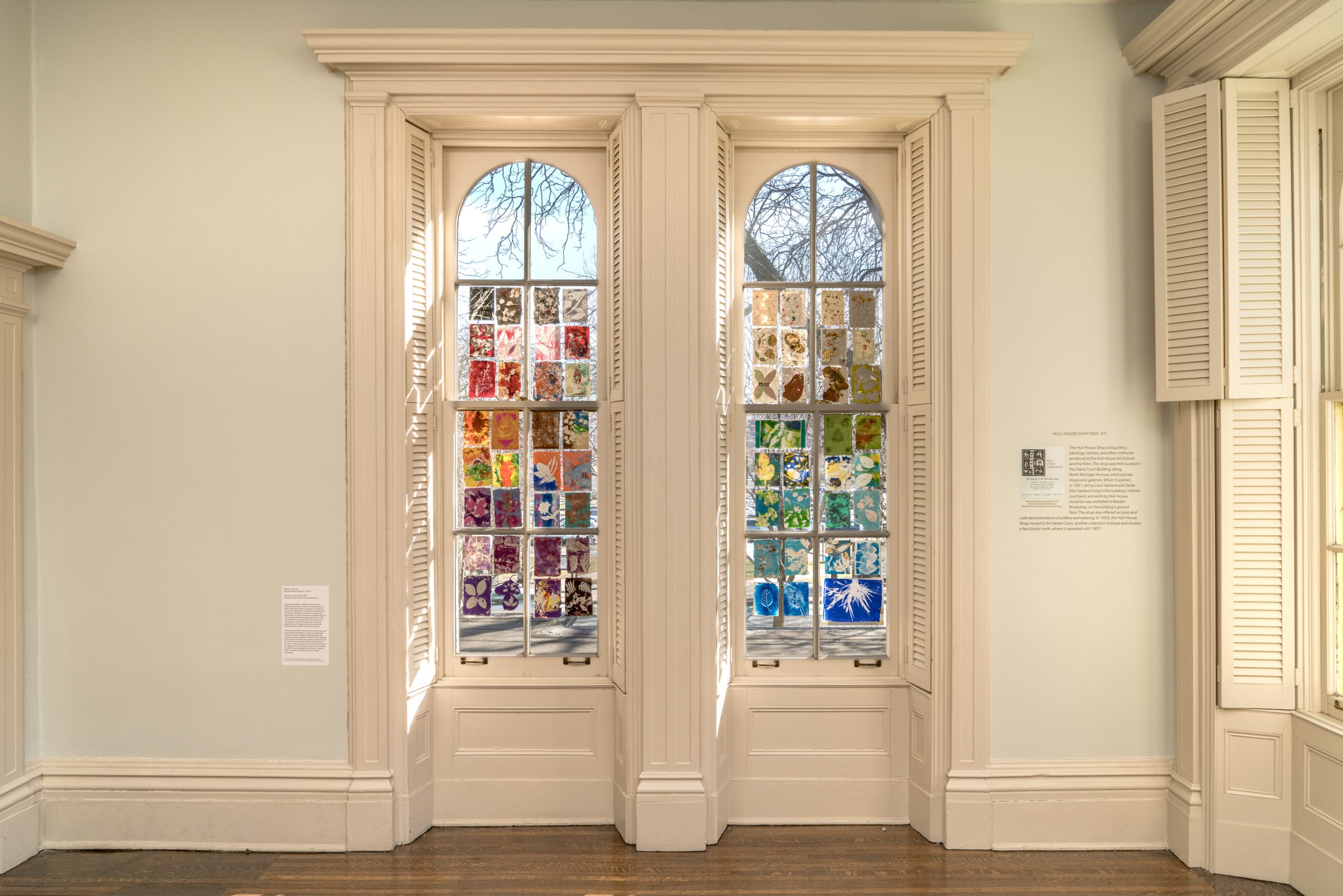 Two tall arched windows with white shutters feature colorful stained glass art panels, with various designs and patterns, allowing sunlight to cast vibrant colors inside a bright gallery room.