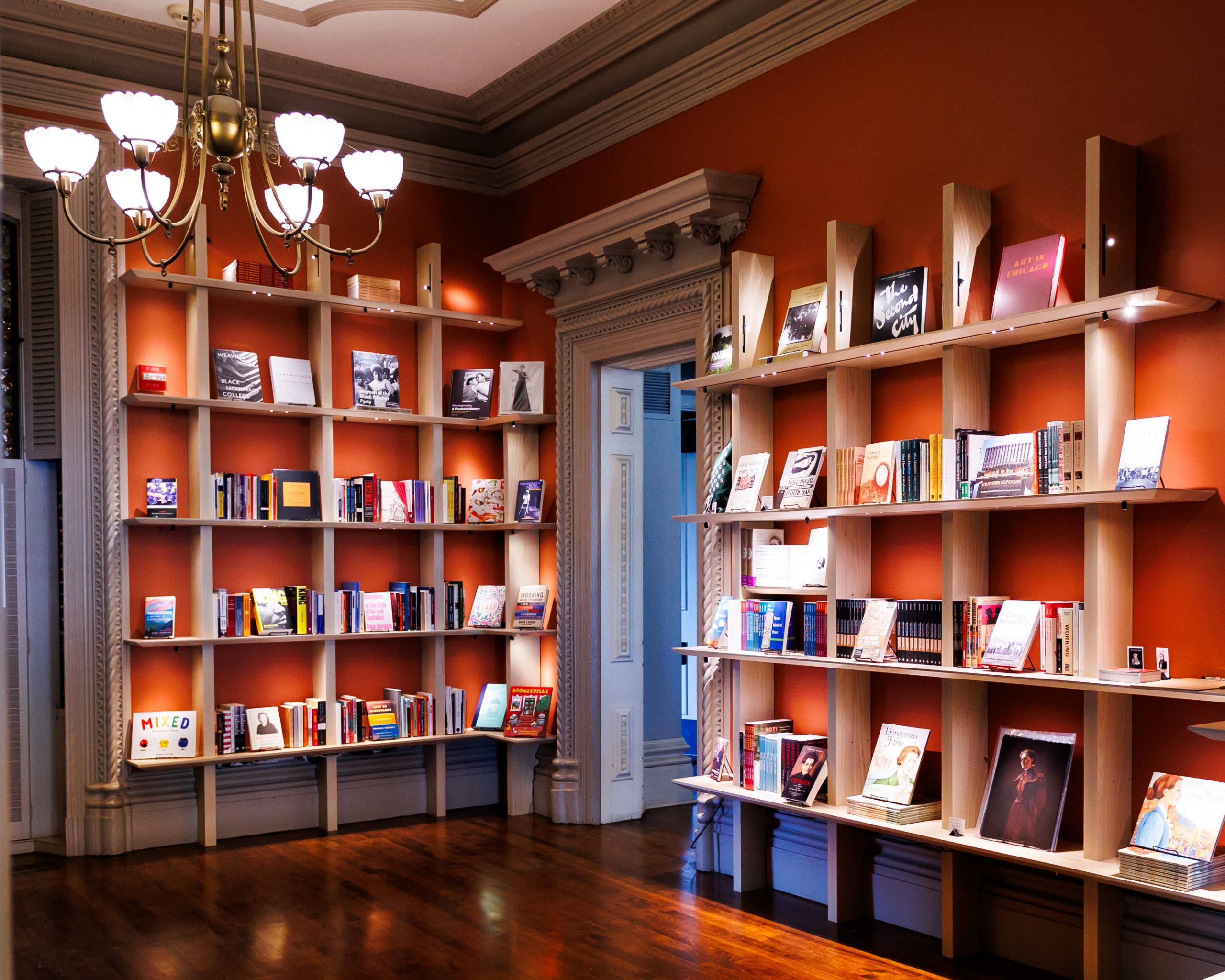 A well-lit room with orange walls featuring two large wooden bookshelves filled with books and magazines. A classic chandelier hangs from the ceiling, and sunlight highlights the polished wooden floor.