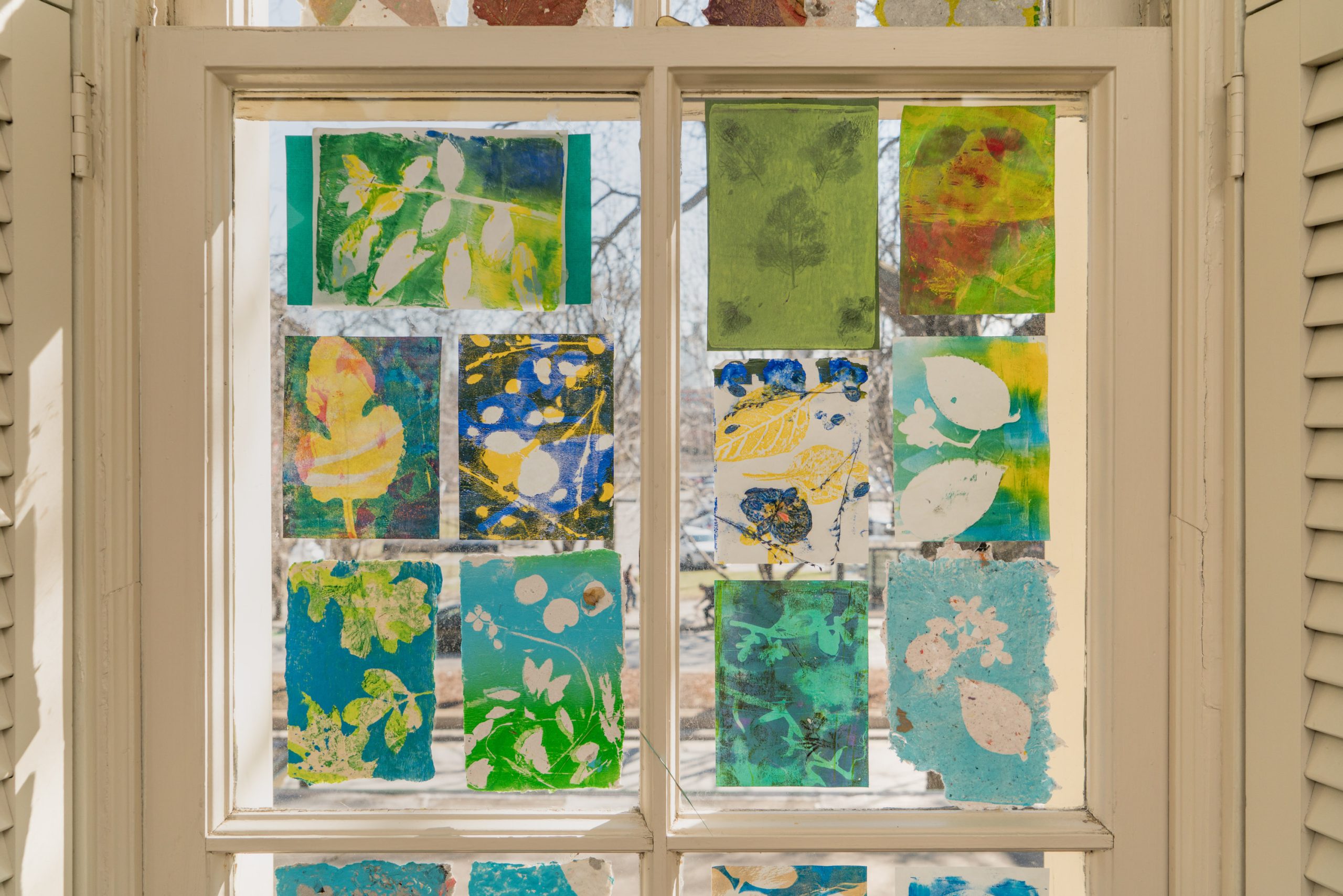 A window with colorful, botanical-themed prints in blue, green, and yellow hues, each featuring leaf and plant designs, is displayed in the glass panes, letting sunlight filter through.