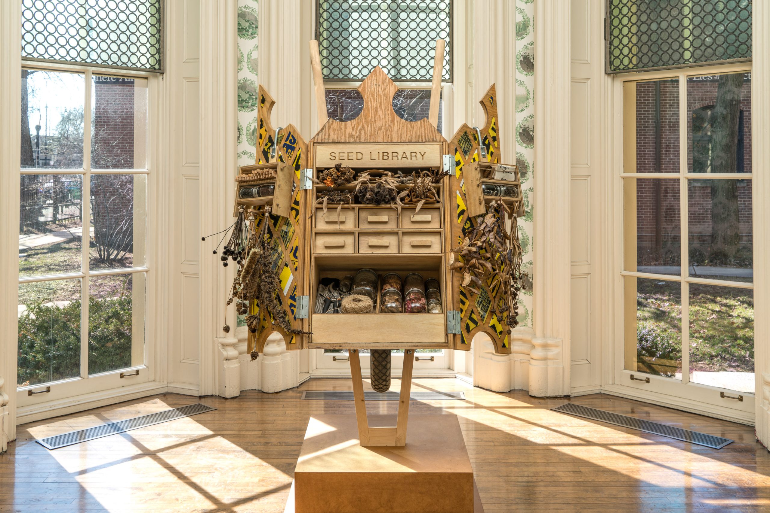 A wooden structure labeled Seed Library stands in front of tall windows, with drawers, seed jars, and dried plants displayed on and around it, set in a bright, ornate room with green patterned window panes.