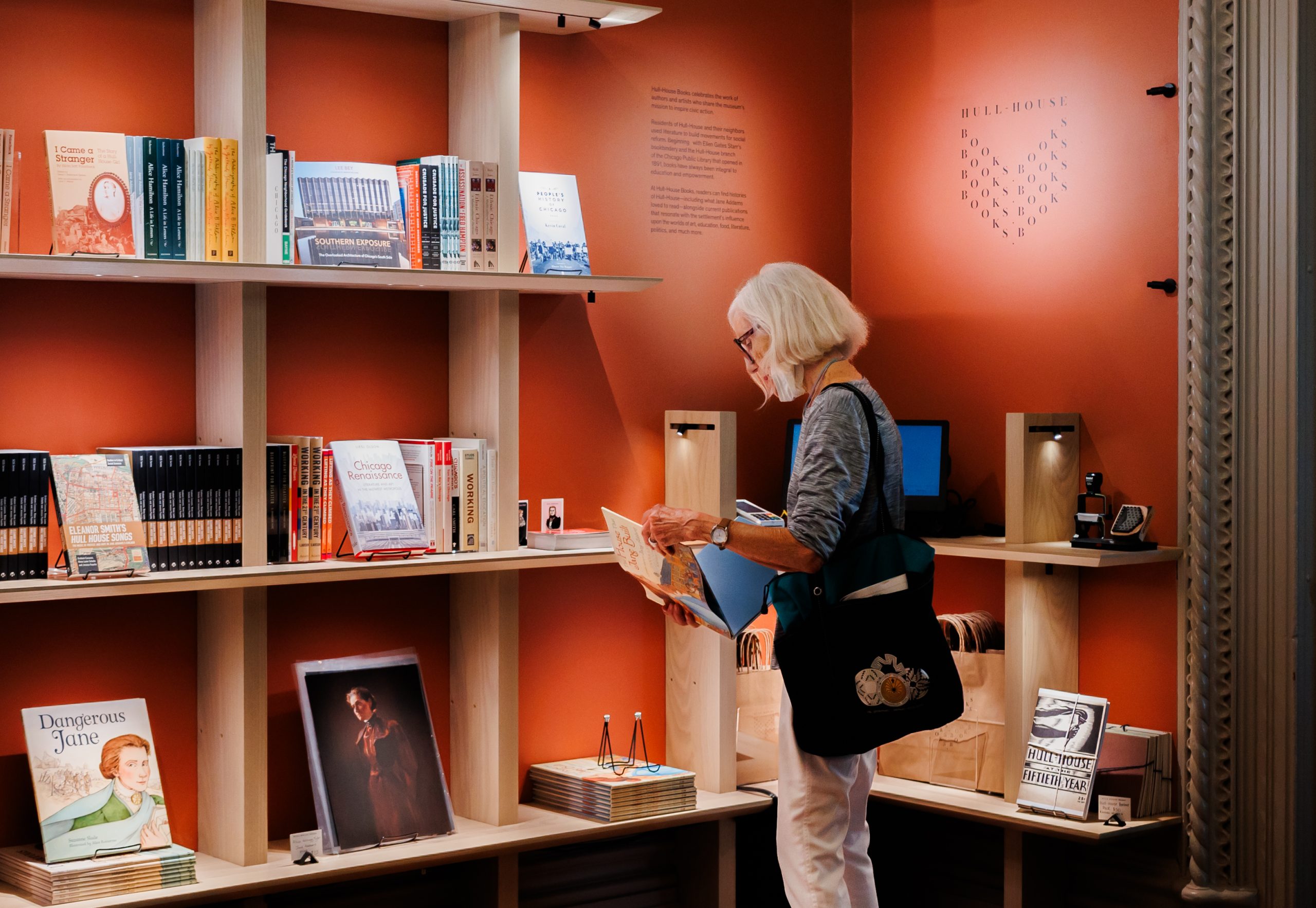 An older woman with white hair reads a book while standing in a cozy bookstore with orange walls and wooden shelves filled with various books and magazines.