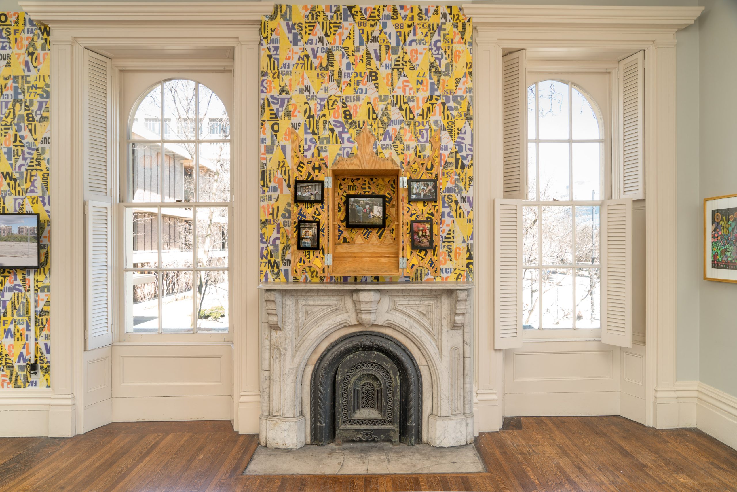A bright room with a white marble fireplace, colorful alphabet-patterned wallpaper, and framed art around a wooden box display above the mantle. Two tall windows with white shutters let in sunlight and show a snowy scene outside.