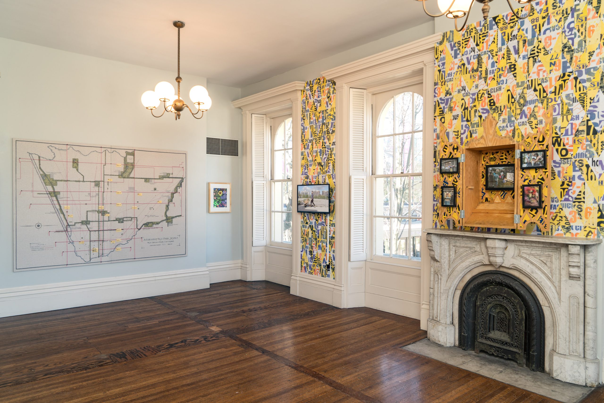 A bright room with wooden floors, a decorative white fireplace, two large windows, colorful letter-patterned wallpaper, framed art on the walls, and modern chandeliers hanging from the ceiling.