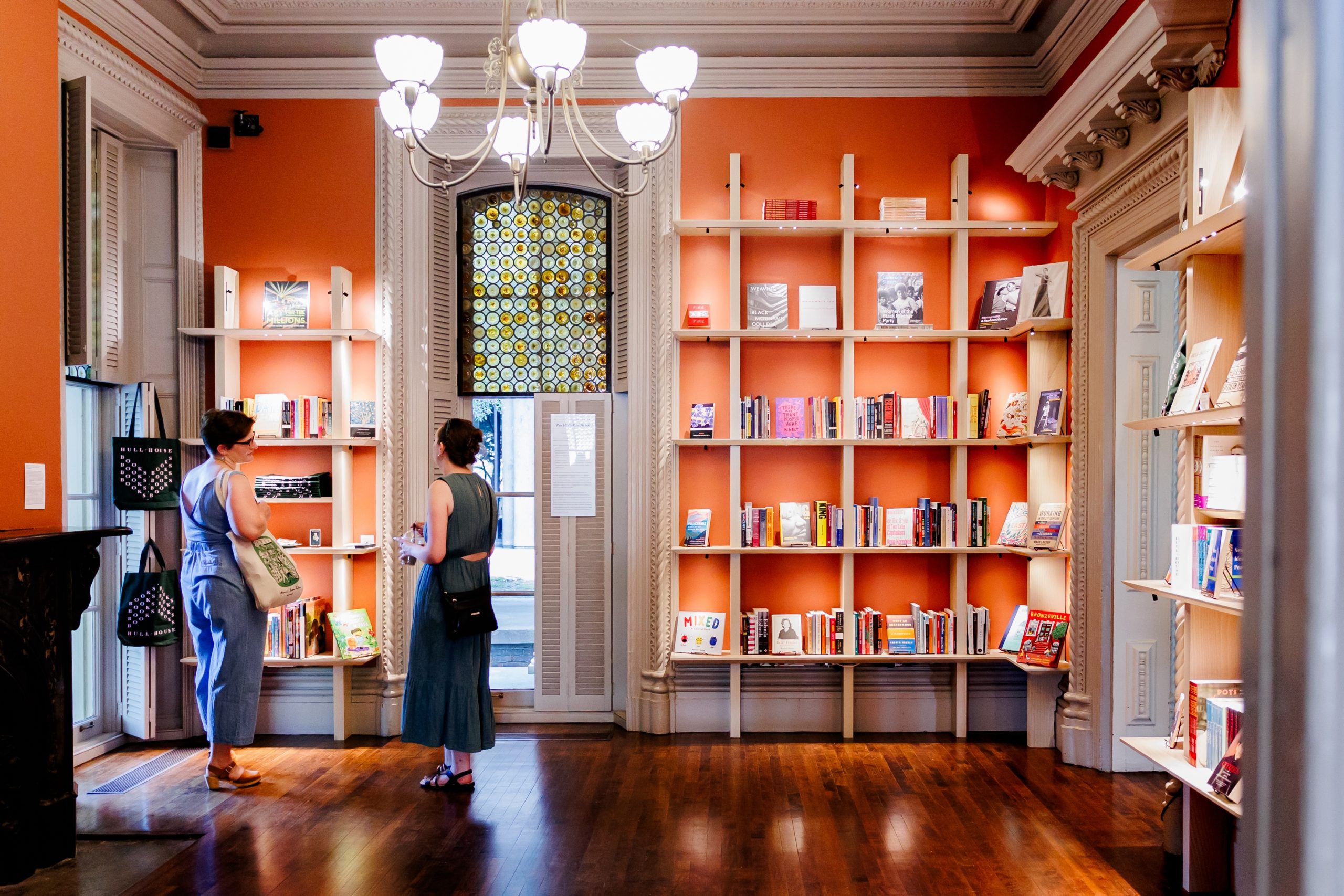 Two women stand in a brightly lit room with orange walls and tall bookshelves filled with books, talking and browsing. A chandelier hangs from the ceiling, and sunlight streams through a decorative window.