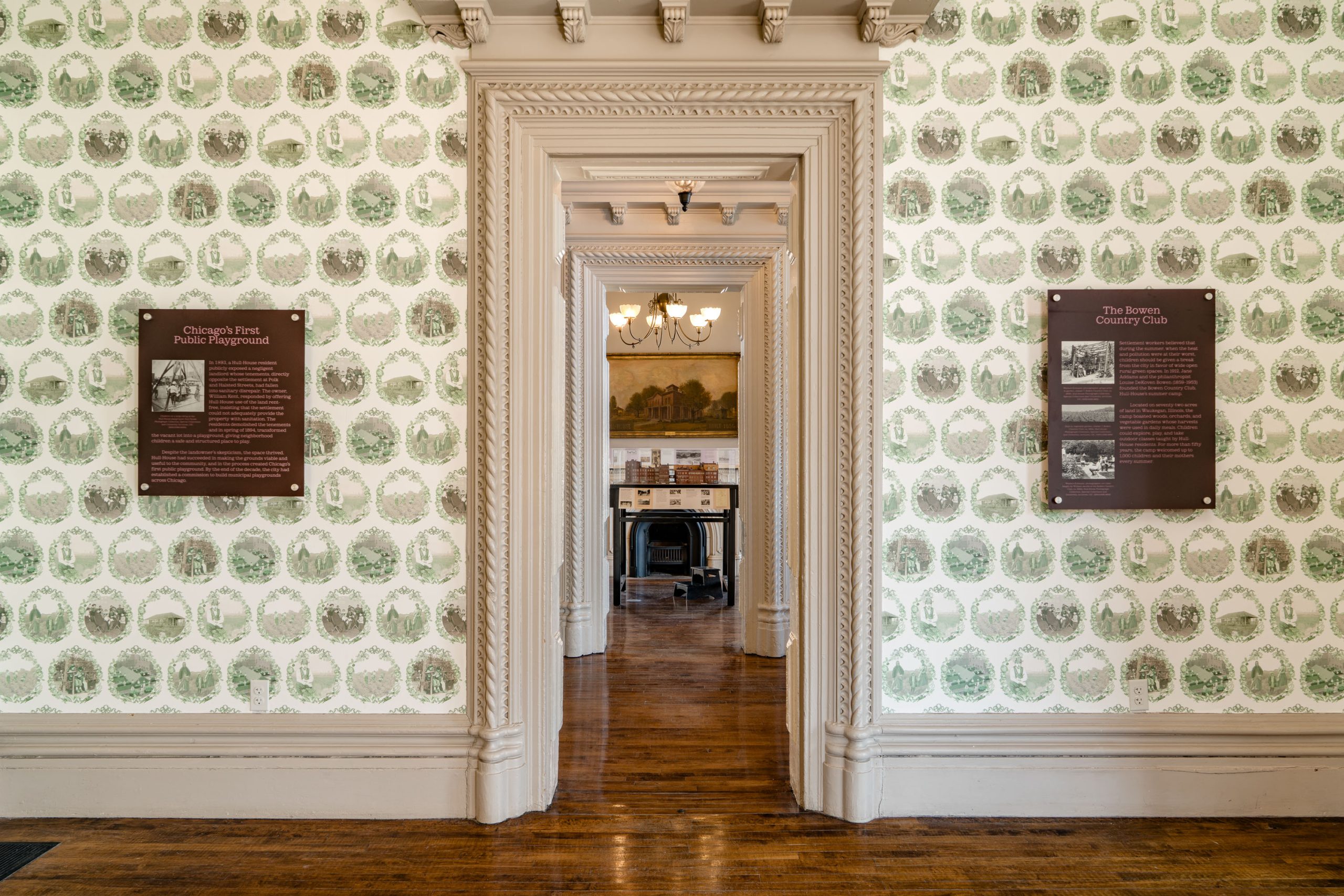 A hallway with ornate trim, patterned wallpaper, and wooden floors. Two informational plaques are mounted on either side of a doorway, leading to a room with a chandelier, painting, chairs, and a desk in the background.