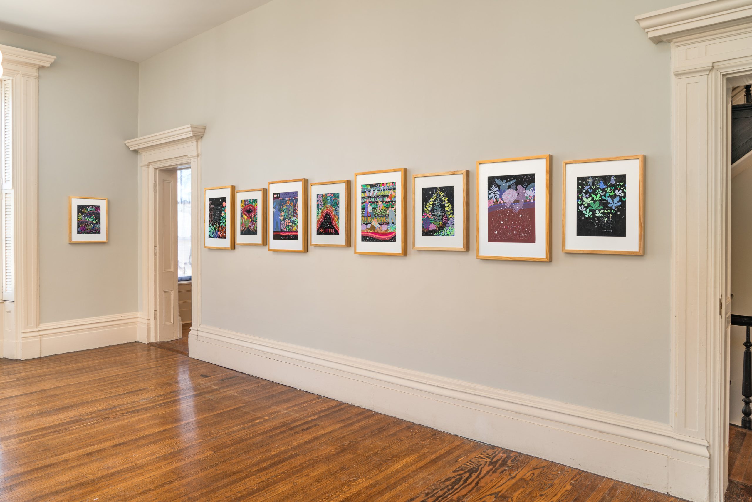 A row of colorful, framed artworks hangs on a light beige wall in a bright room with hardwood floors and decorative white trim around doors and windows.
