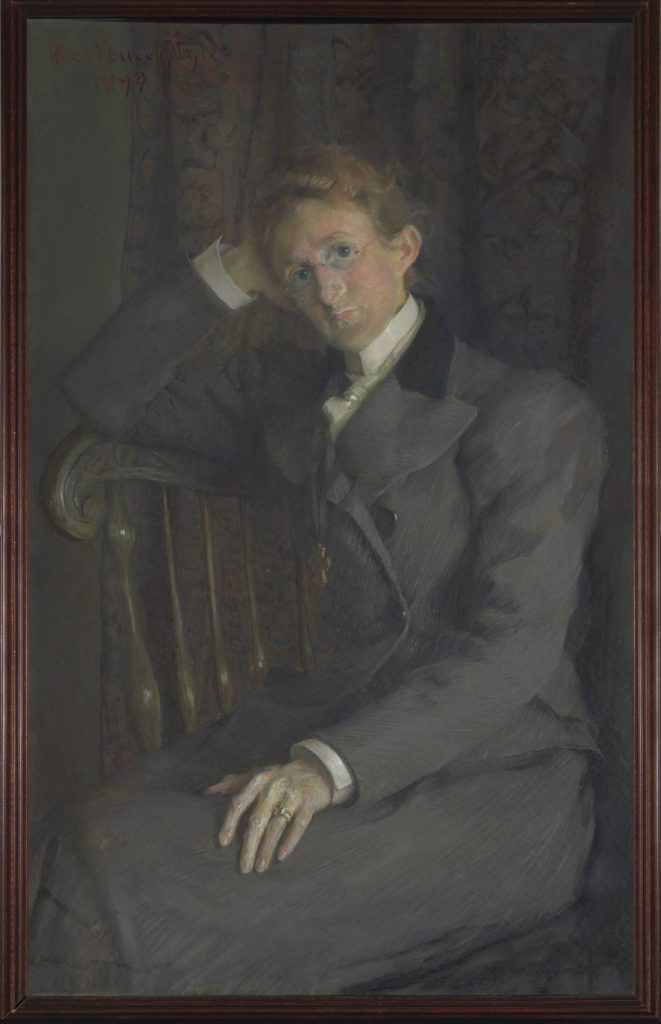 A person with short, light hair wearing round glasses and a gray suit sits on a wooden chair, resting one arm on the backrest and gazing thoughtfully forward. The background features a dark, patterned curtain.