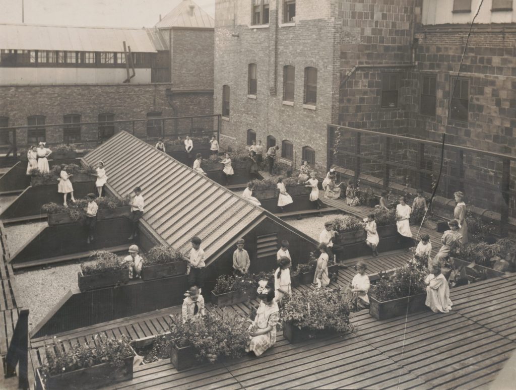 Black-and-white photo of women and children sitting and standing in a rooftop garden, surrounded by planters and brick buildings, with some people tending to plants or conversing.