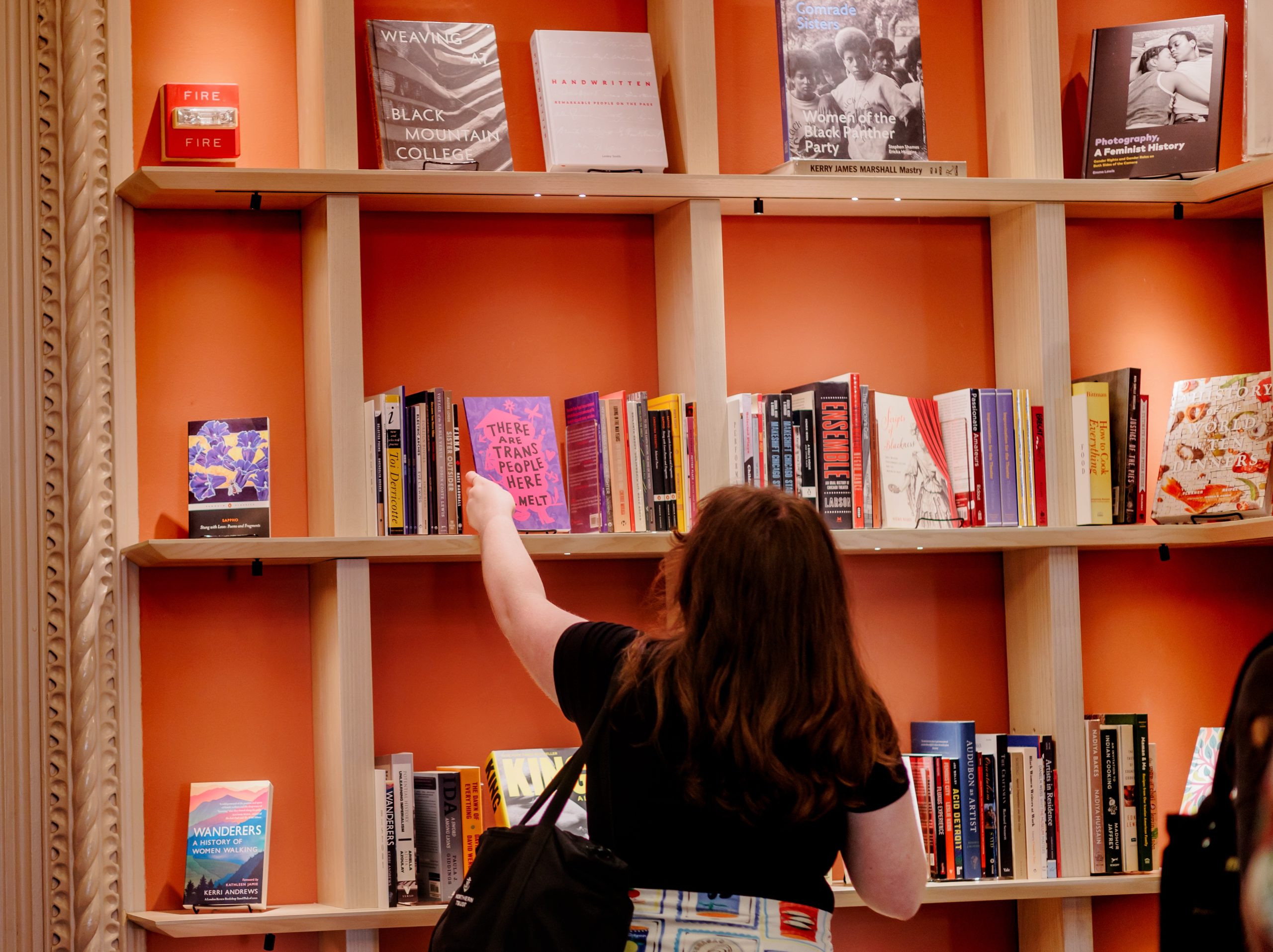A woman with long brown hair reaches for a book on a wooden bookshelf filled with colorful books against an orange wall.