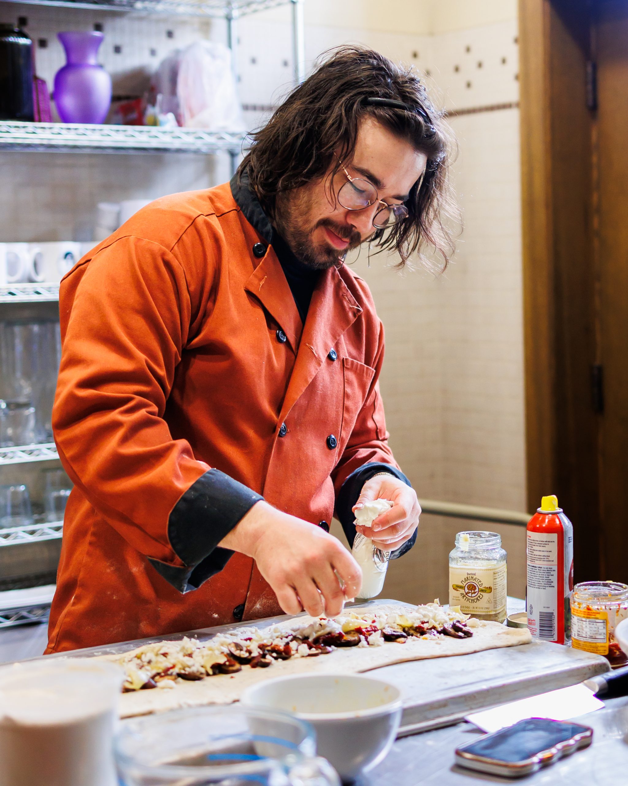 A person wearing an orange chef coat is smiling while preparing food, sprinkling cheese or another topping onto a long pastry in a kitchen with shelves of dishes and ingredients in the background.