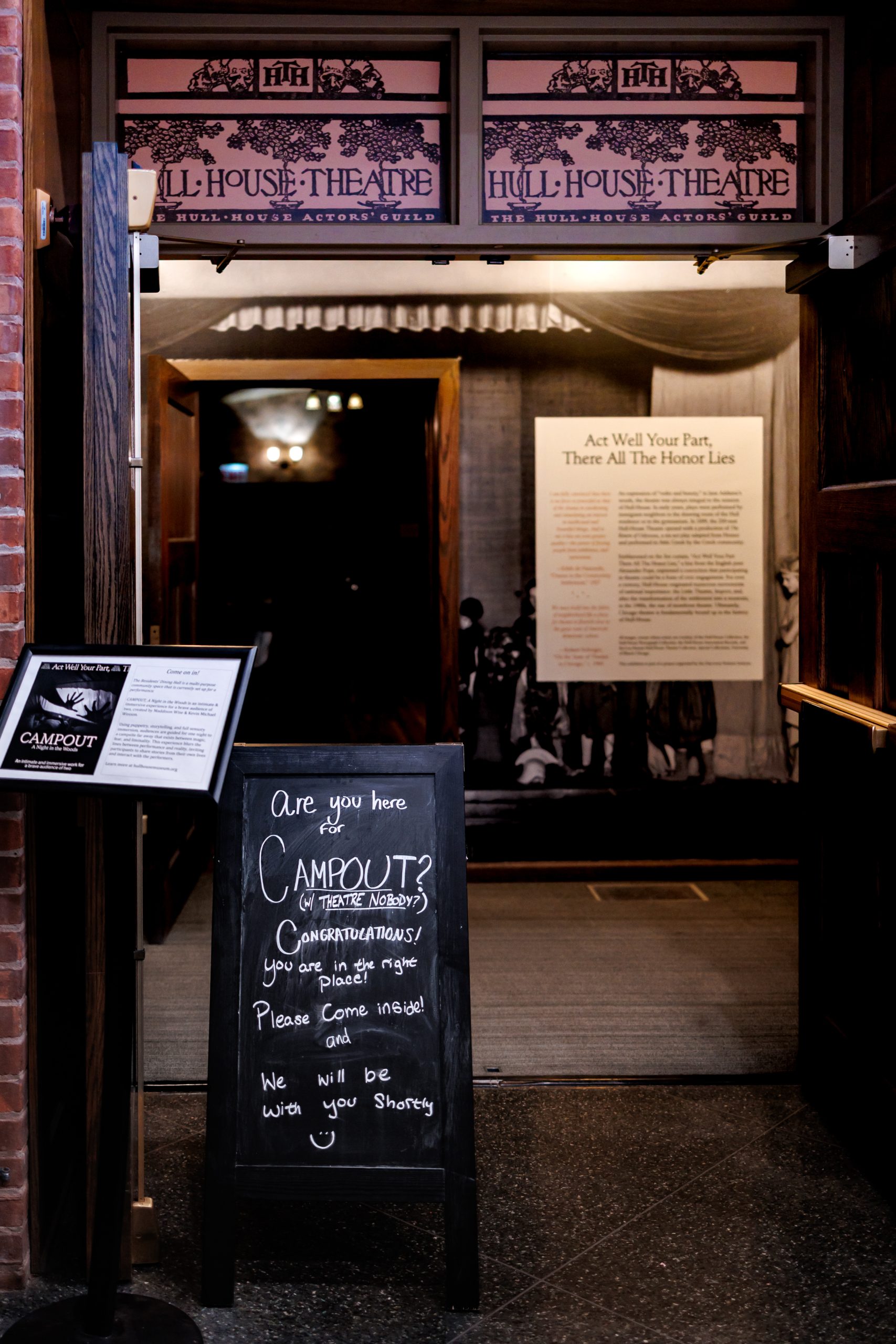 Entrance to Hull-House Theatre with doors open, a blackboard sign inviting Campout! guests inside, and an informational display visible in the background. Warm lighting creates a welcoming atmosphere.