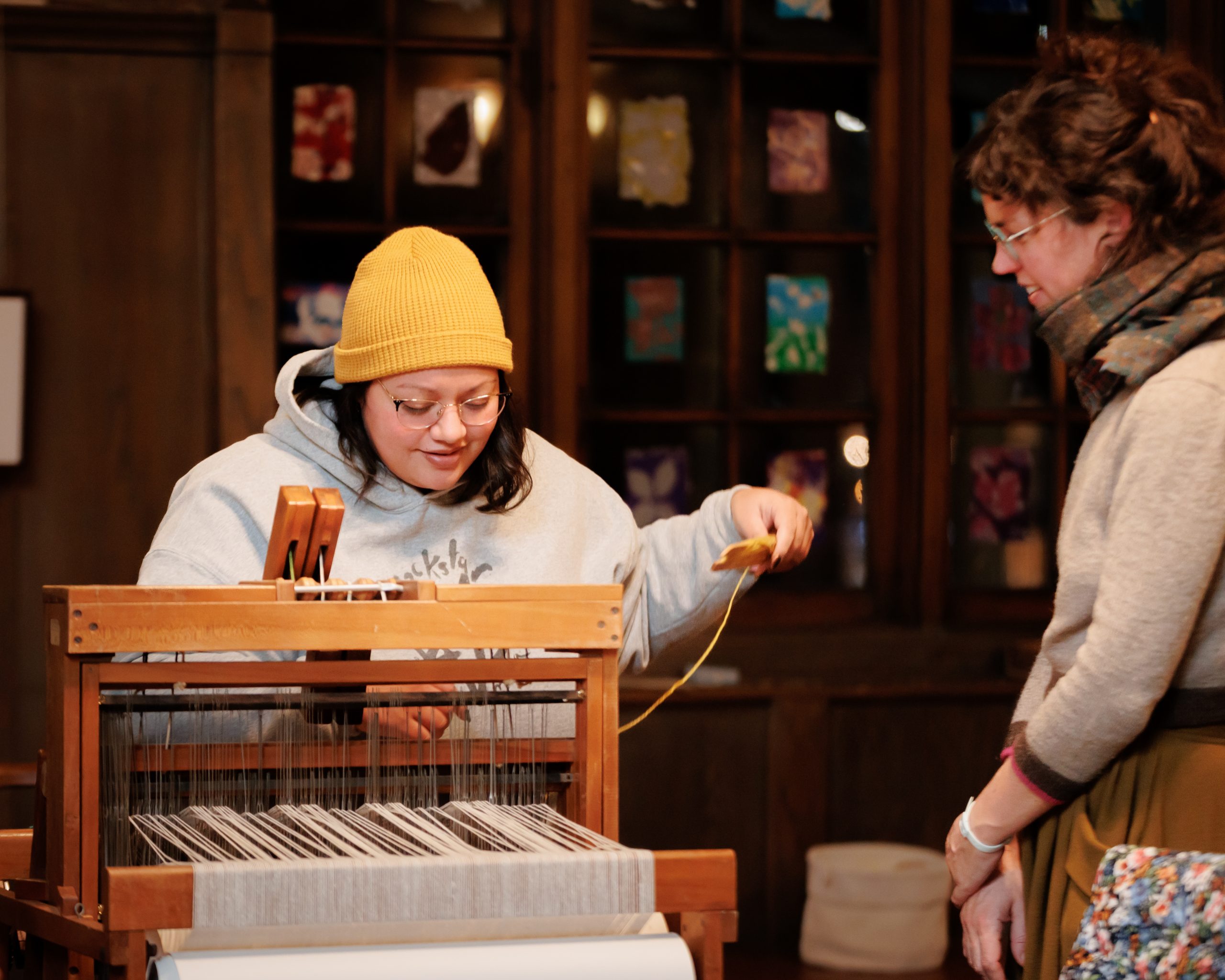 Two people are standing by a wooden loom. One, wearing a yellow beanie and gray hoodie, weaves thread while the other watches. Colorful artwork is displayed on a windowed wall behind them.