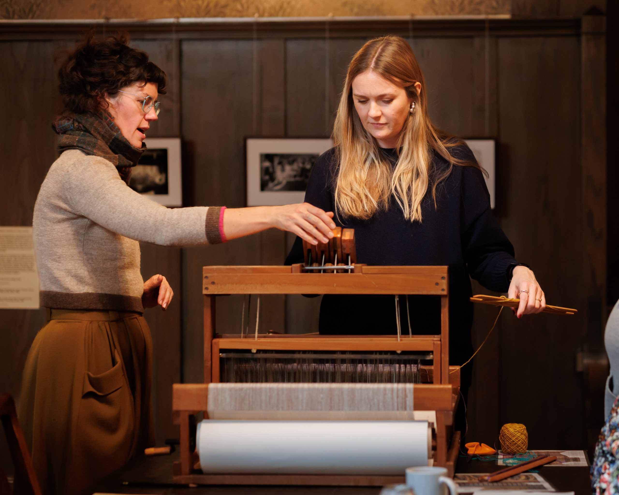 Two women stand at a wooden loom, one demonstrating how to use it while the other observes and holds yarn. They are indoors, with framed photos on the wall in the background.