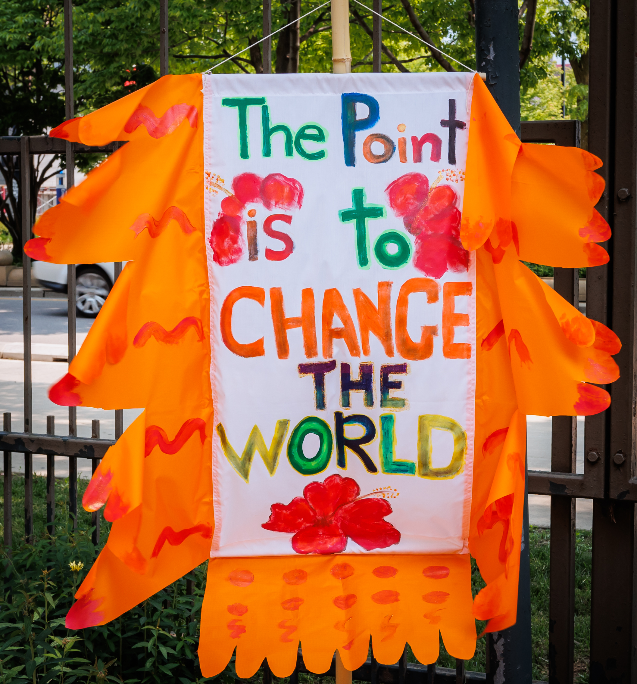 A colorful banner with orange fringes reads, The point is to change the world in bright, multicolored letters, decorated with red flowers. The banner hangs outdoors near a metal fence and greenery.