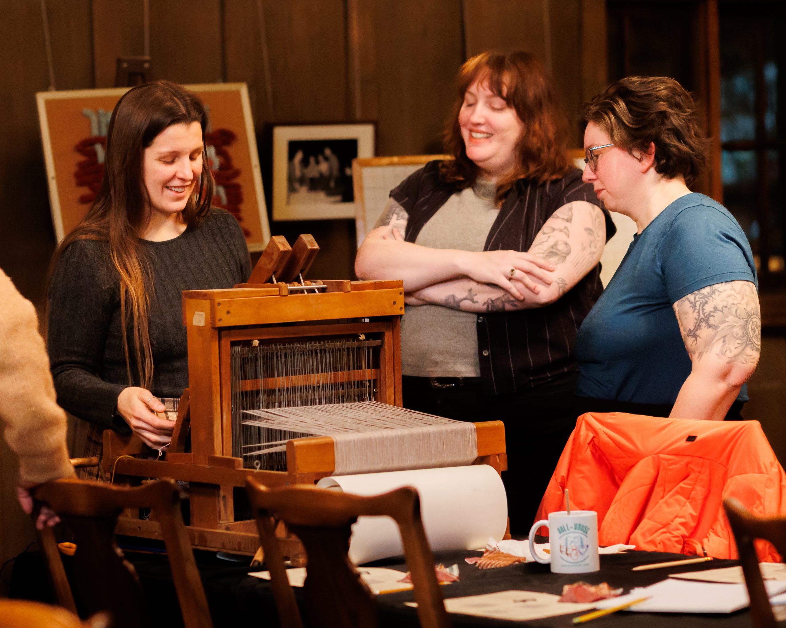 Three women smile and converse around a small wooden loom at a table indoors. Papers, a mug, and an orange jacket are on the table. Framed artwork decorates the wooden-paneled wall behind them.