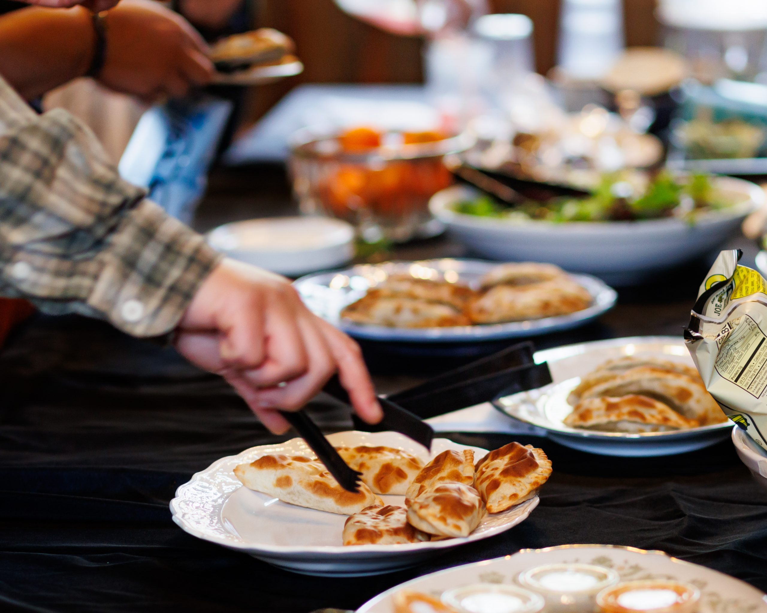 A person uses tongs to serve flatbread onto a plate from a buffet table filled with various dishes, including salads and dipping sauces. Other hands are also reaching for food.