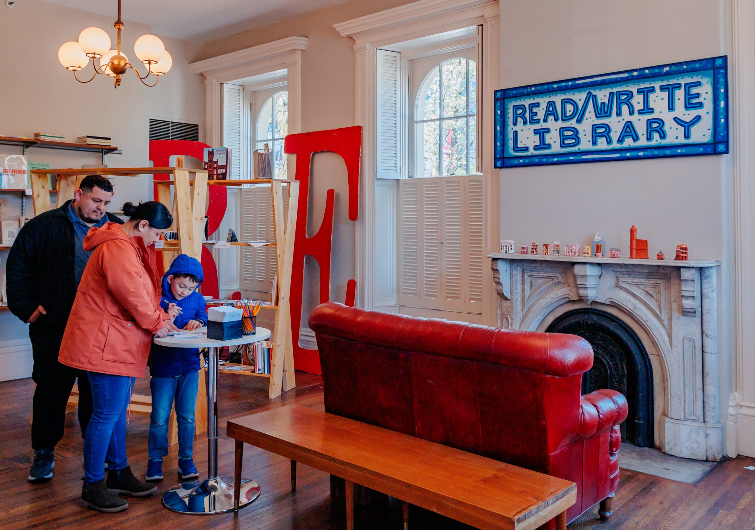A family stands around a small table in a cozy library with large windows, bookshelves, and a red couch. A sign reads Read/Write Library above a decorative fireplace.