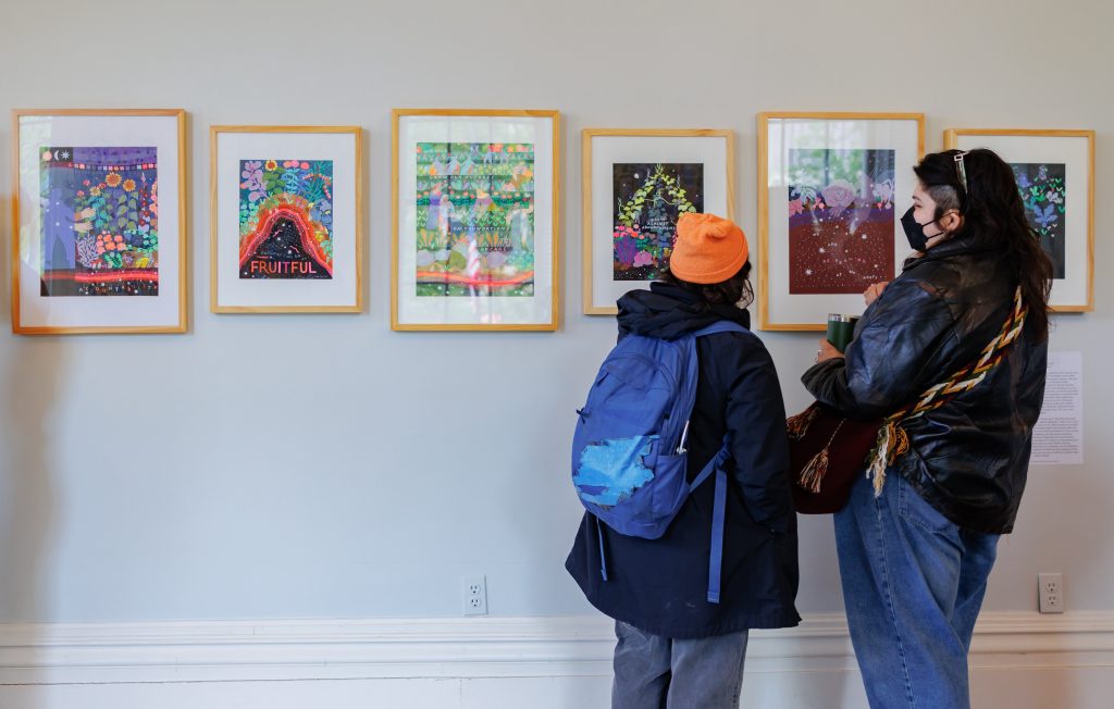 Two people stand in front of a wall displaying six colorful, framed artworks. One person wears a blue jacket and orange beanie, the other wears a mask and holds a cup. Both are viewing the art closely.