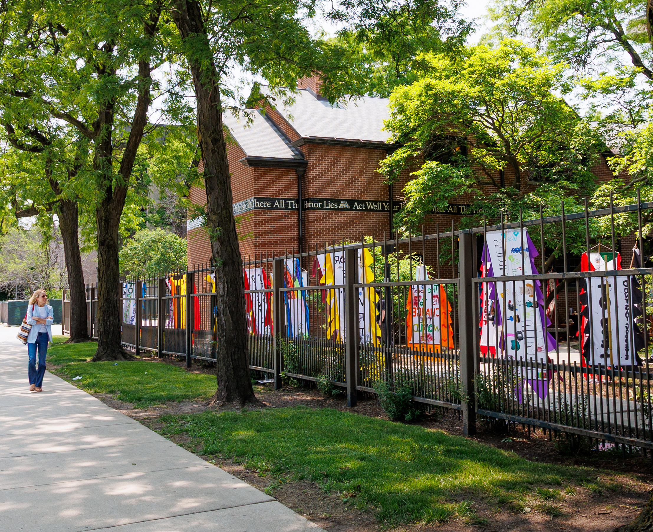 A woman walks on a sidewalk beside a fence decorated with colorful vertical banners, in front of a brick building surrounded by leafy green trees on a sunny day.