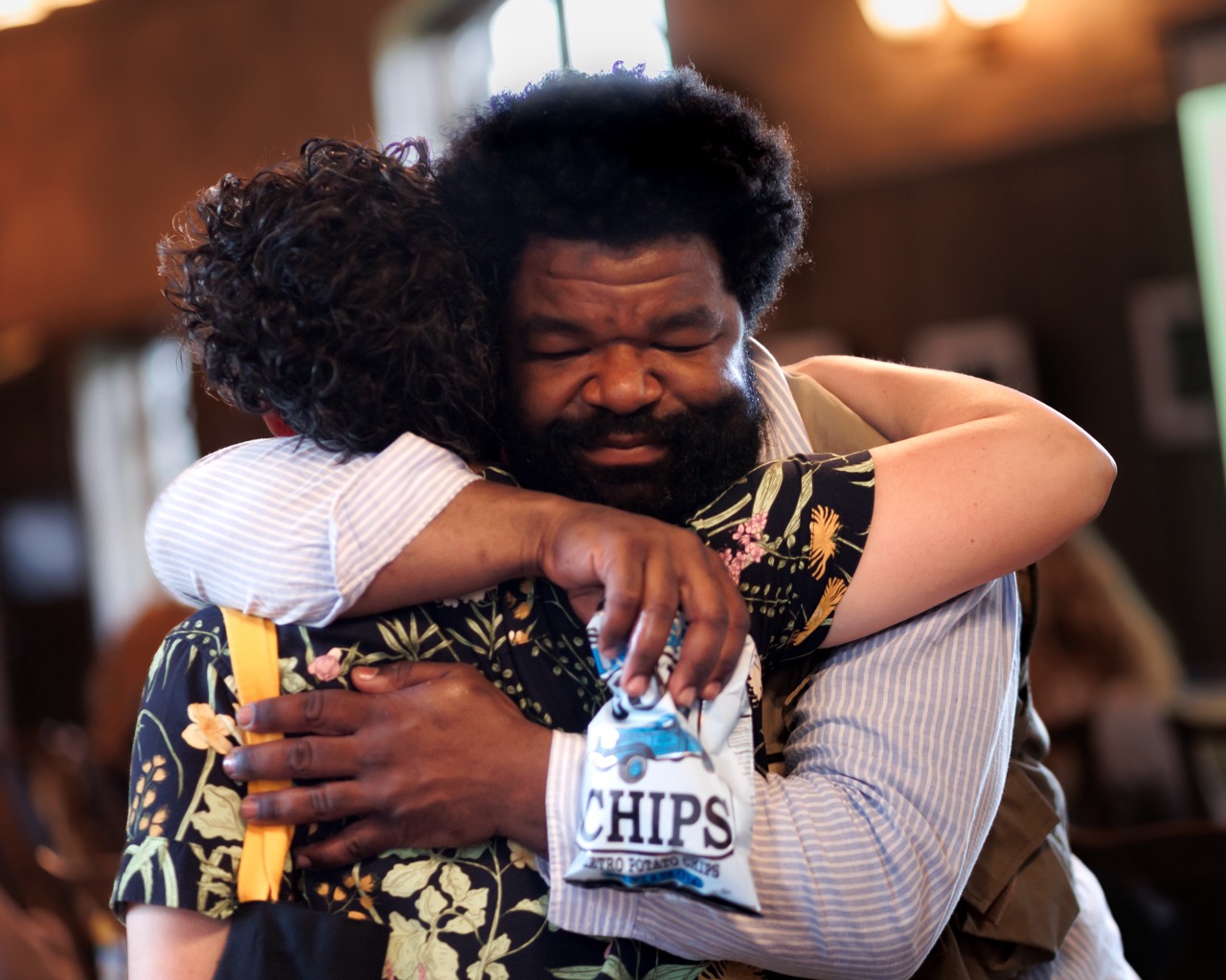 Two people warmly hug indoors. One person, smiling with eyes closed, holds a bag of potato chips. The background is softly lit and out of focus, creating a cozy, intimate atmosphere.