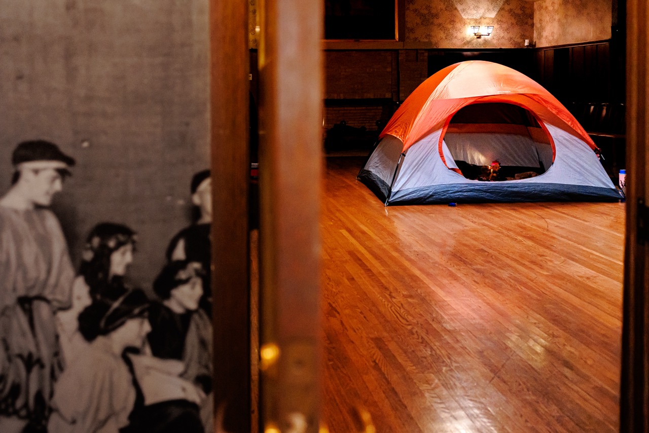 A red and gray tent is set up inside a spacious wooden-floored room, seen through a doorway with a blurred black-and-white photo of people on the left side of the doorframe.