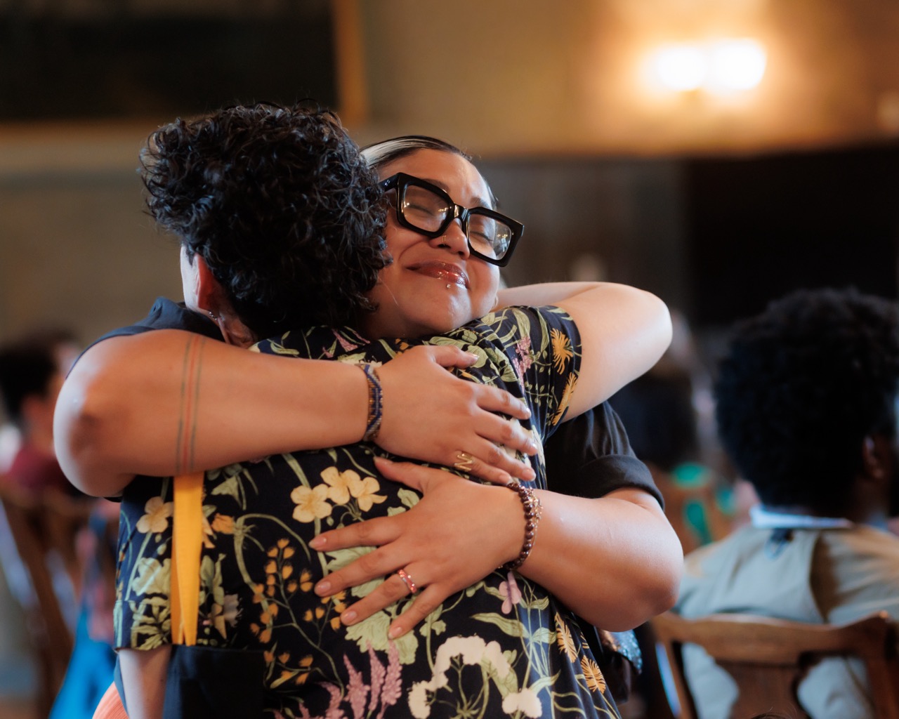 Two people embrace warmly in a hug, one smiling with closed eyes and wearing large glasses. The setting appears to be a warmly lit room, with others seated and out of focus in the background.