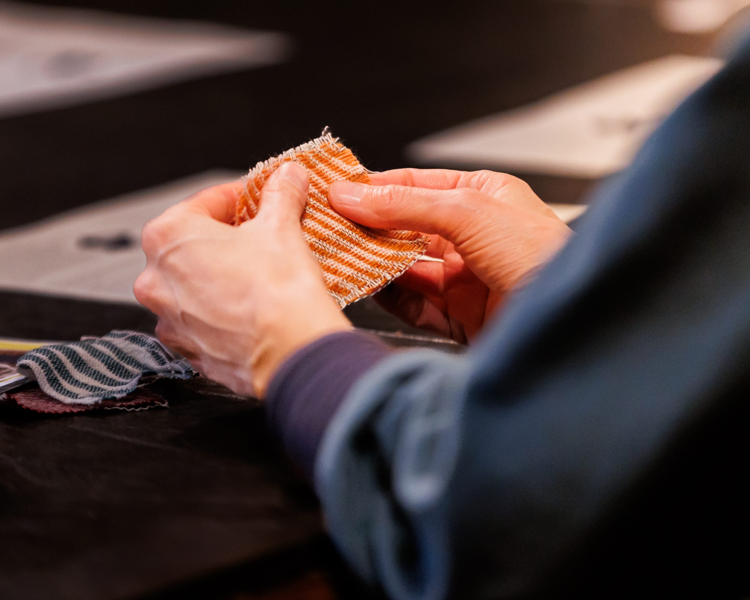A person holding and examining a small, orange-and-white striped fabric swatch with both hands, with other fabric swatches and papers visible on a dark table in the background.