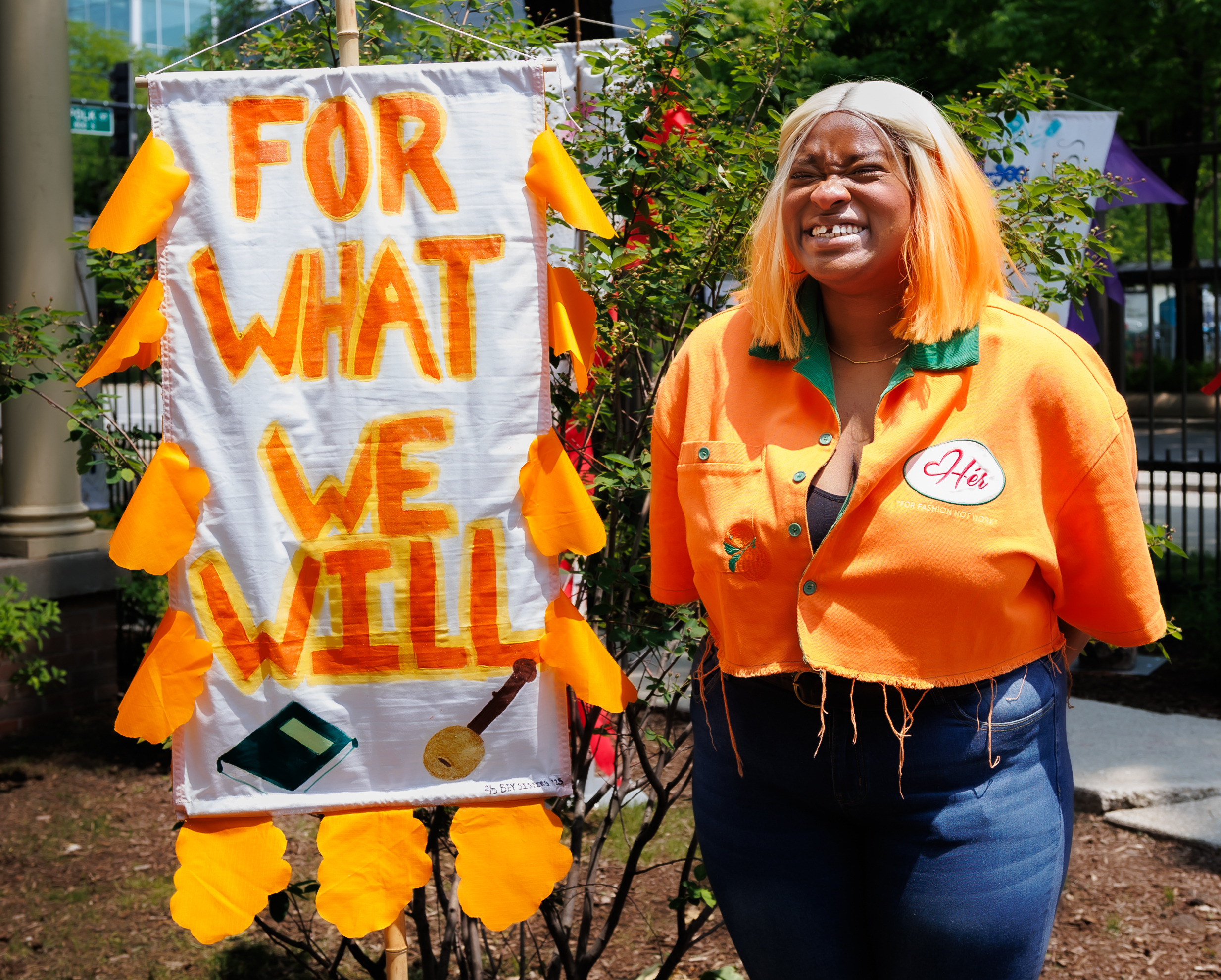 A person with orange hair and an orange shirt stands outside next to a handmade sign that reads FOR WHAT WE WILL, decorated with yellow leaves and drawings of a phone and a key. The person is smiling and squinting.