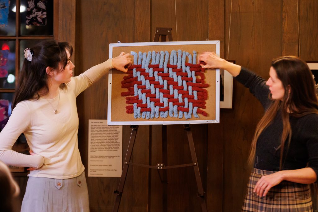 Two women stand beside an easel, pointing at a corkboard displaying a red and blue woven textile pattern, in a warmly lit room with wooden walls.