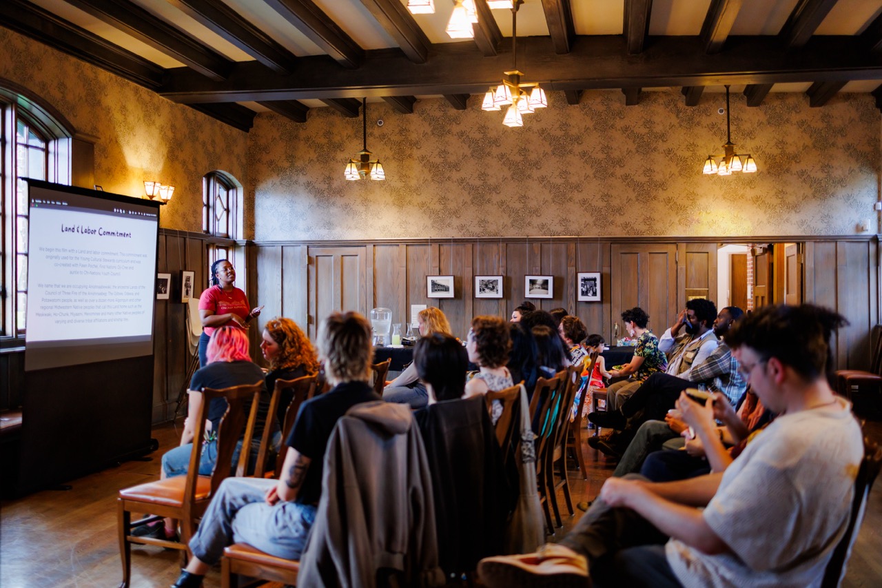 A group of people sit on wooden chairs in a vintage-style room, attentively watching a speaker present slides on a screen at the front. The room has wooden paneling, large windows, and warm lighting.