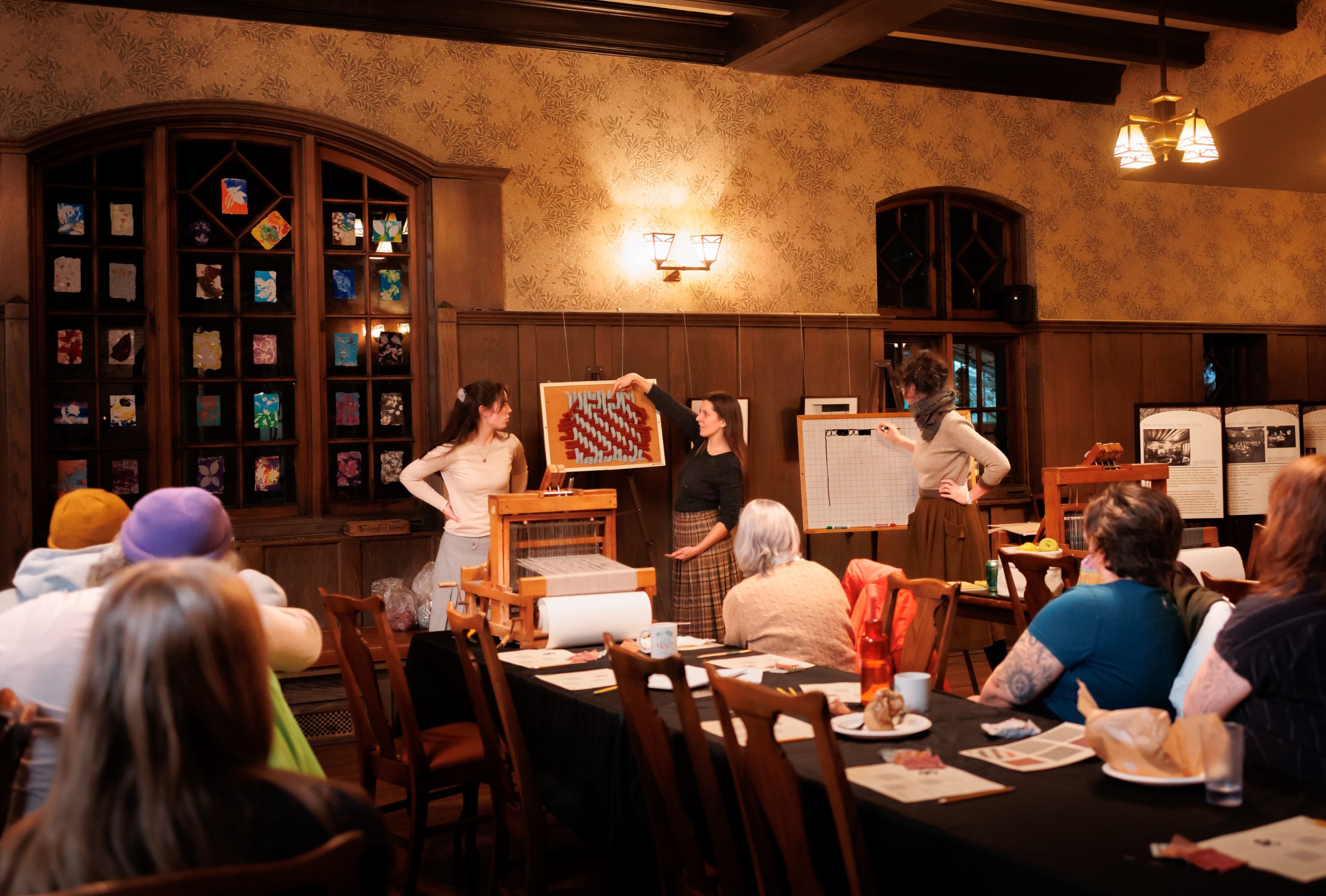 Three women stand at the front of a room holding up a framed art piece to an audience seated at tables. The room has wooden paneling, arched windows, and displays on easels. Warm lighting sets a cozy atmosphere.