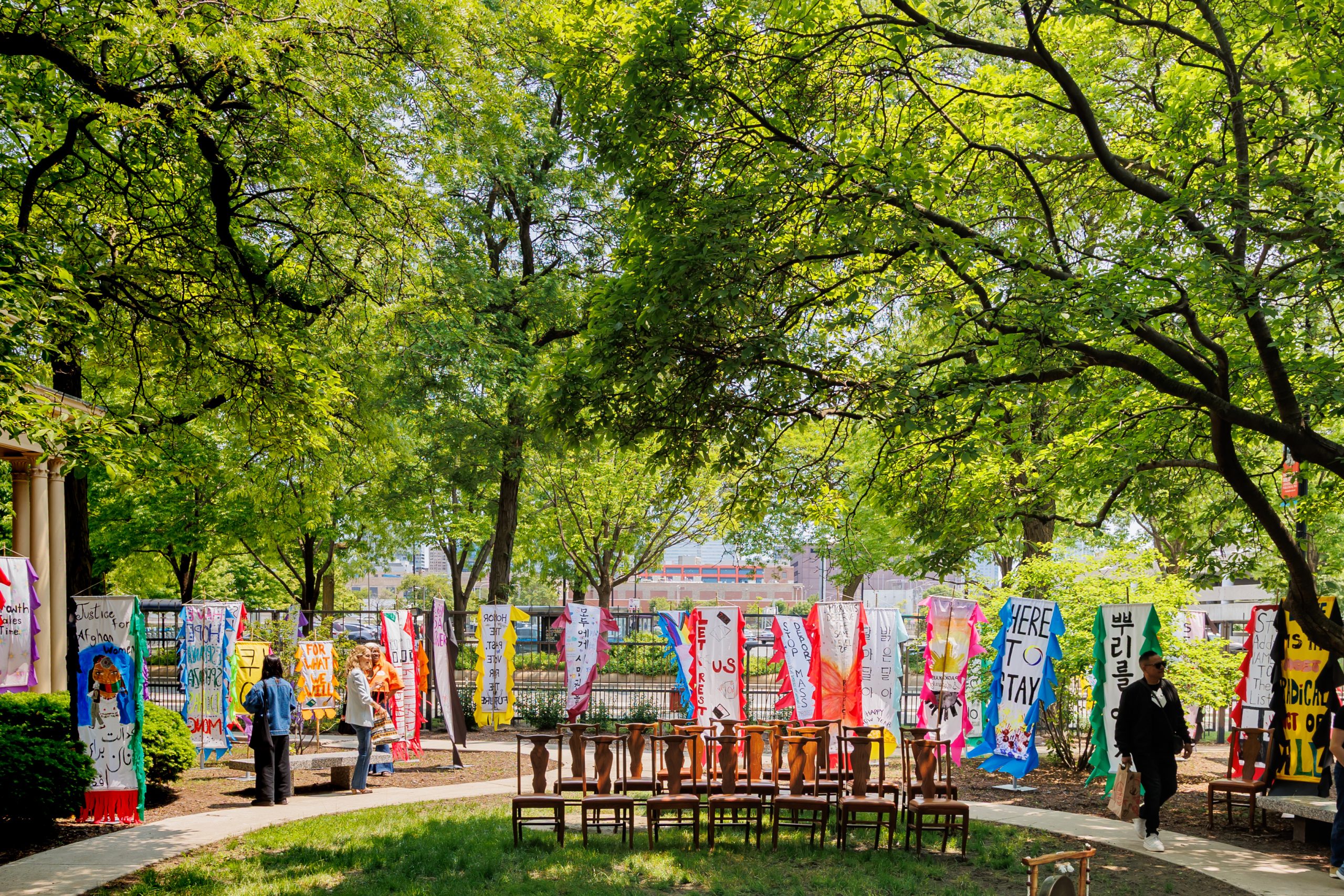 A park scene with many colorful banners hanging from a fence, surrounded by green trees. Several people are walking or standing, and empty wooden chairs are arranged in a circle on the path.