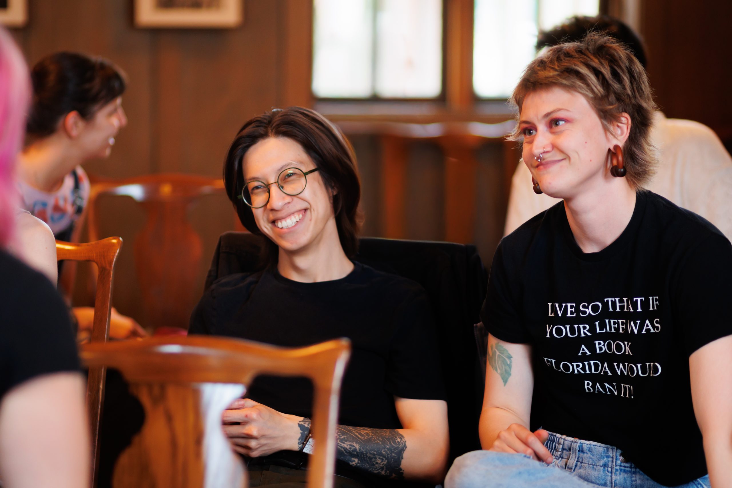 Two people sit smiling in a warmly lit room. One wears glasses and a dark shirt, the other has short hair, earrings, and a black T-shirt with white text. Other people are visible in the background, engaged in conversation.