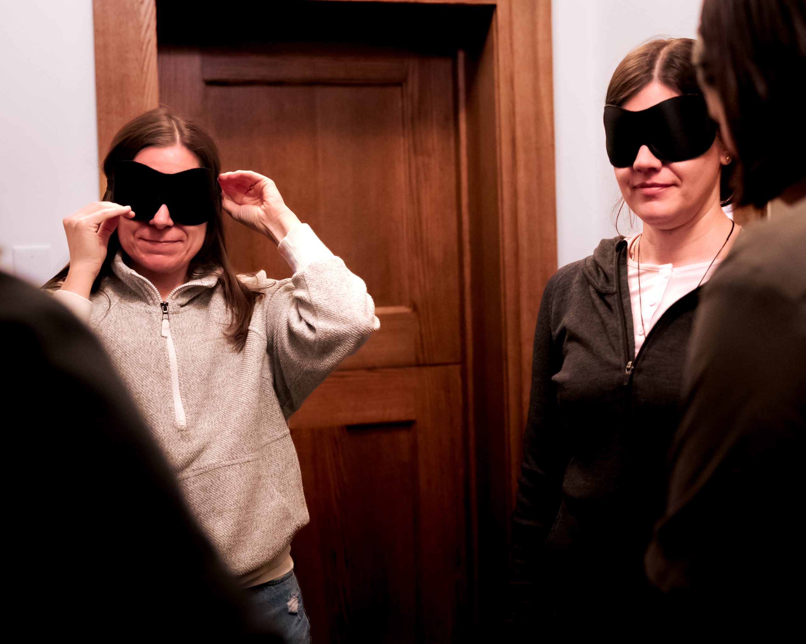 Two women in casual clothing stand indoors, both wearing black blindfolds. One woman adjusts her blindfold while the other stands facing her. A closed wooden door is visible in the background.