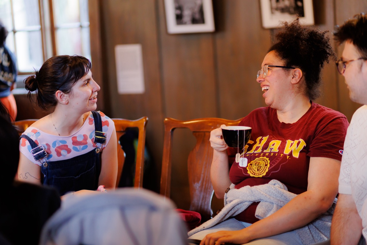 Two people sit and chat warmly indoors; one is holding a mug and laughing, wearing glasses and a red shirt, while the other listens, smiling, dressed in overalls with a colorful shirt. Framed photos hang on the wall behind them.