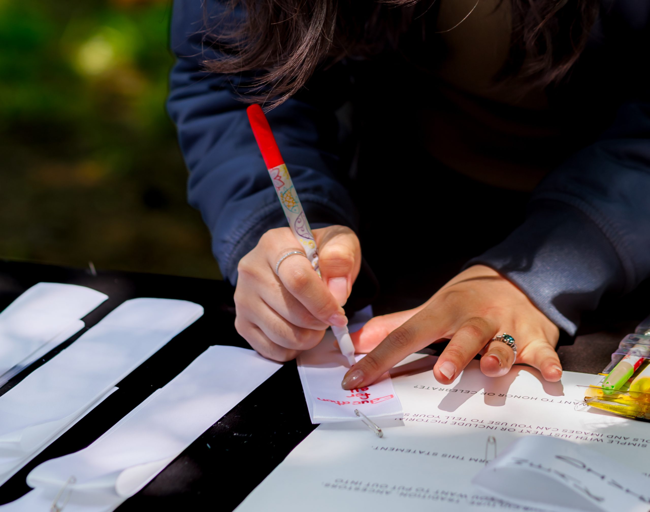 A person writes on a white paper strip with a red-tipped pen at an outdoor table, surrounded by other paper strips and documents. Only their hands and part of their upper body are visible.
