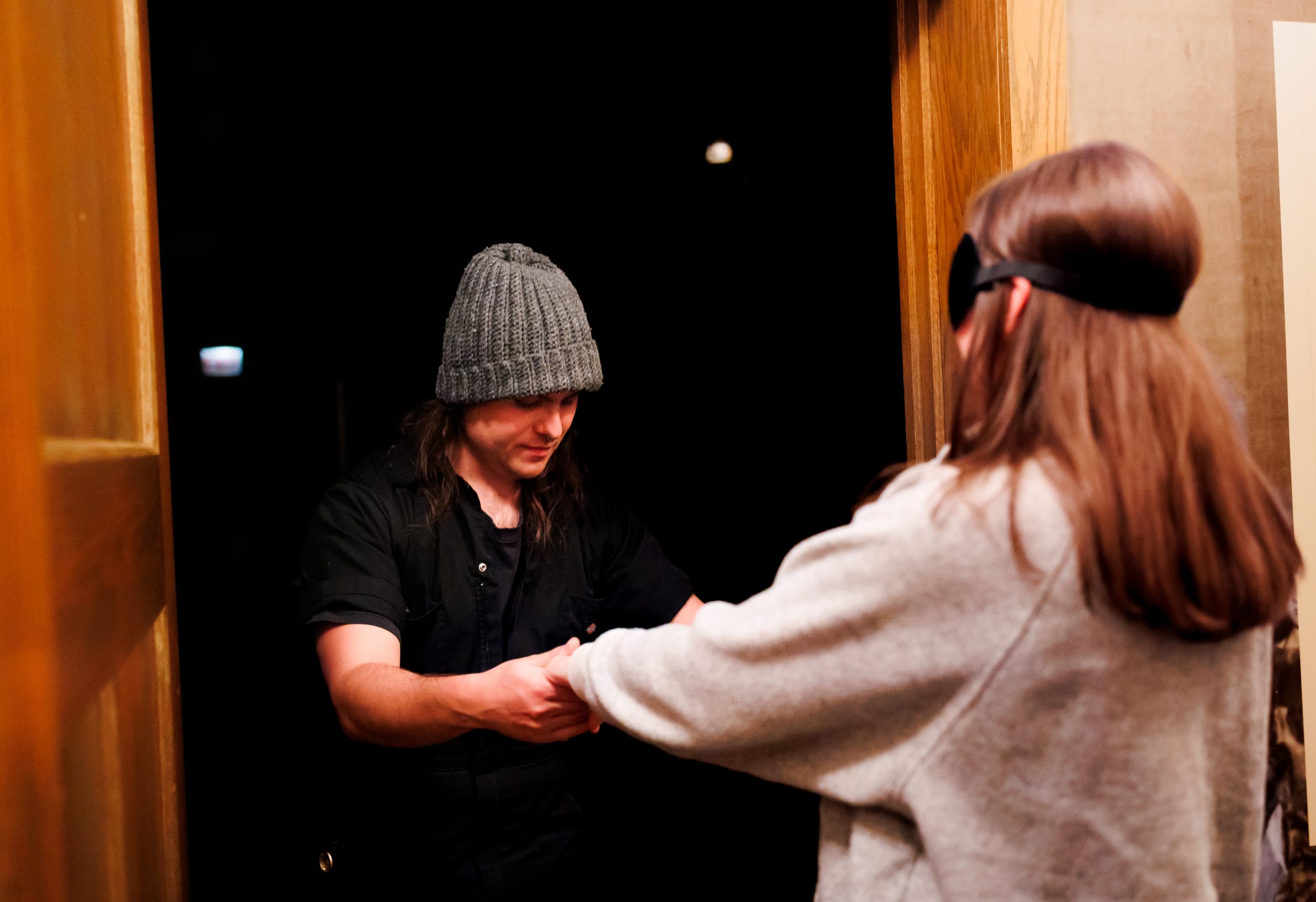 A person wearing a knit beanie stands in a doorway, holding hands with another person in a sweatshirt and blindfold. The scene appears to take place at night.