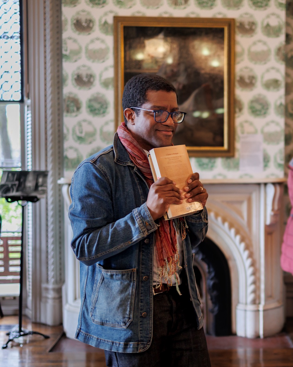 A person wearing glasses, a scarf, and a denim jacket stands indoors, smiling and holding a book to their chest. Behind them is a decorative fireplace and a framed painting on a patterned wall.