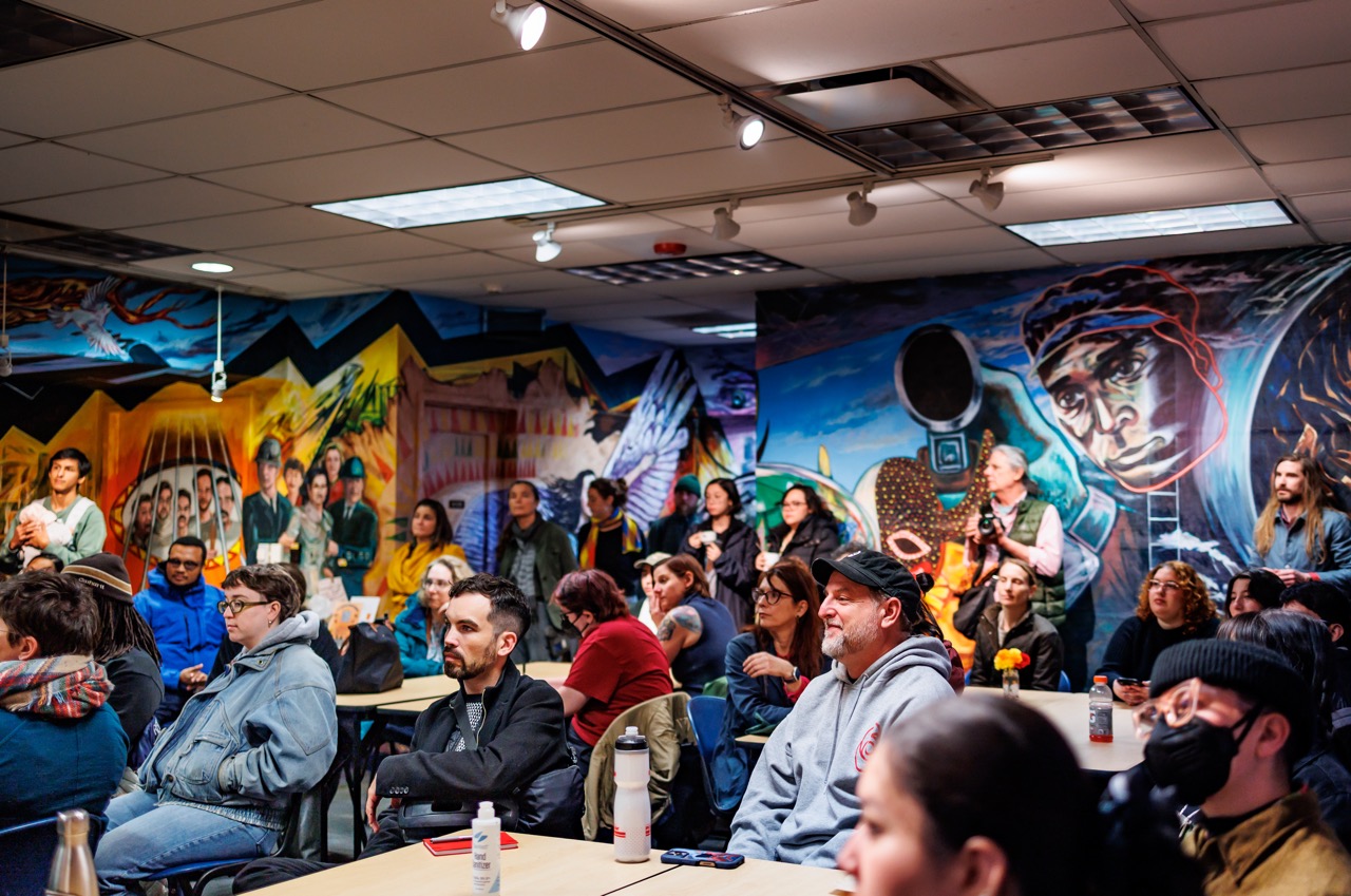 A diverse group of people sit and stand attentively in a vibrant, mural-filled room, engaging in a community event or meeting. Tables and drinks are visible, creating a casual, welcoming atmosphere.