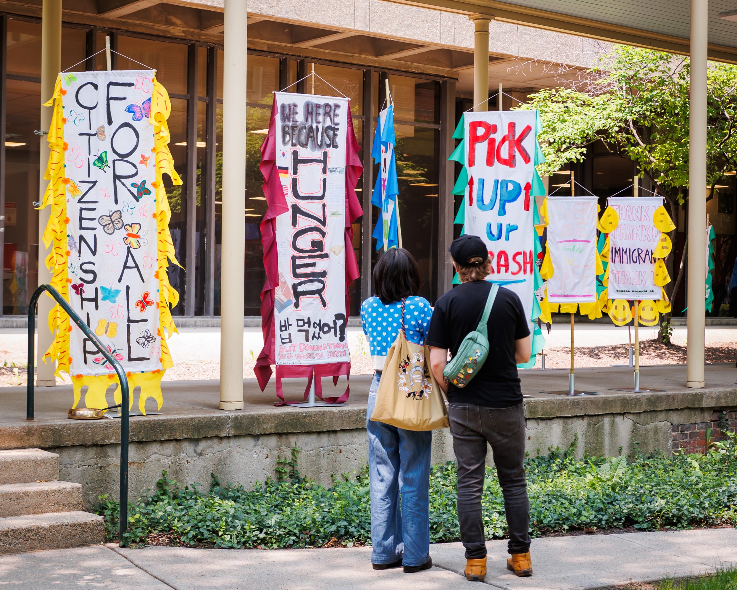 Two people stand outside, looking at colorful banners with messages like “For All,” “We’re Here Because of Hunger,” “Pick Up Your Trash,” and “Immigrant,” displayed on a building’s walkway.