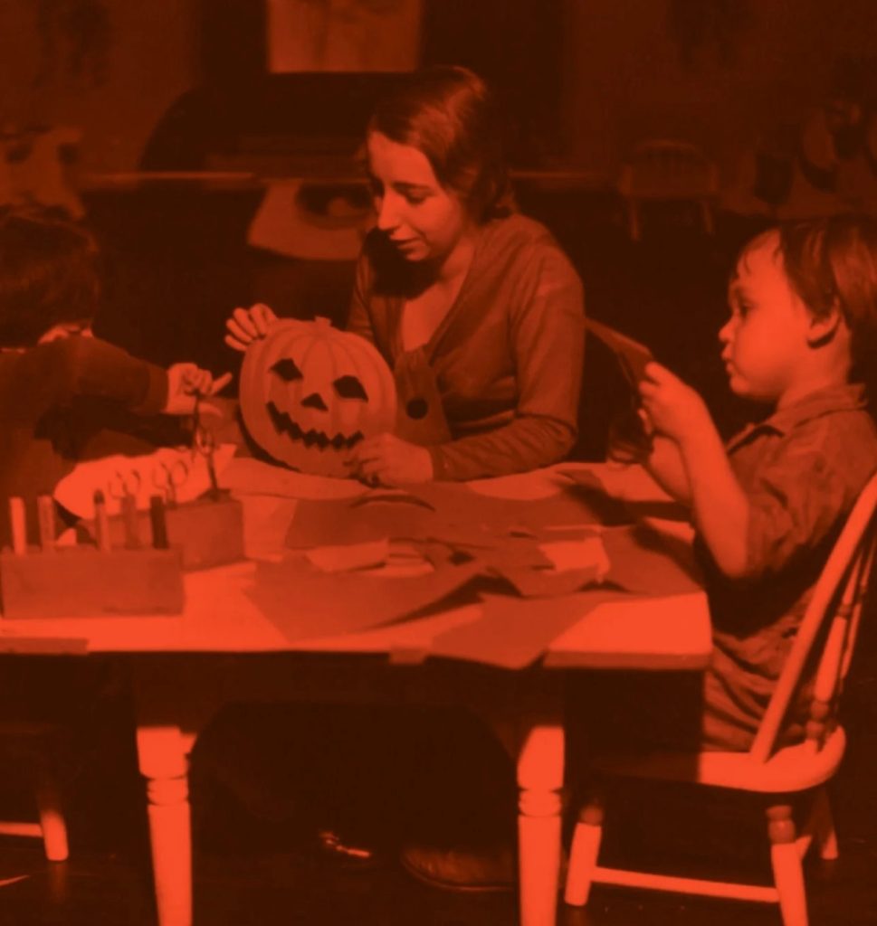 Three children sit at a table doing Halloween crafts, including carving a jack-o’-lantern and cutting out paper shapes. The scene has a reddish tint, and the table is scattered with supplies.