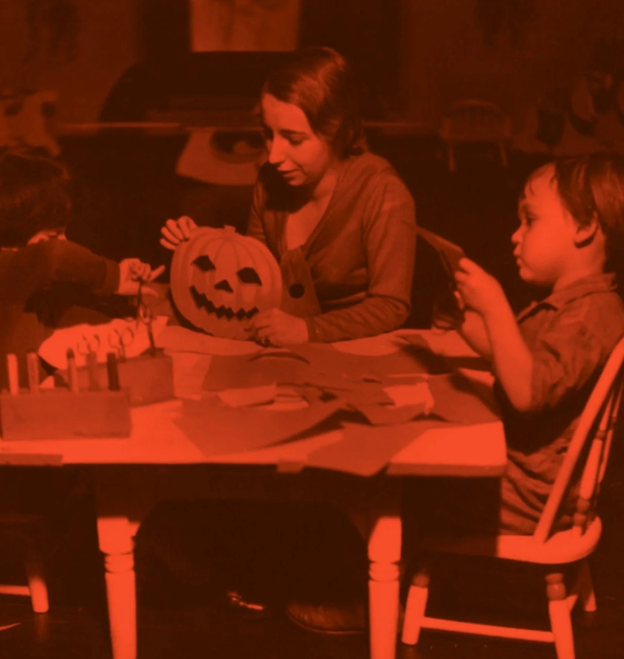 Three children sit at a table doing Halloween crafts, including carving a jack-o’-lantern and cutting out paper shapes. The scene has a reddish tint, and the table is scattered with supplies.