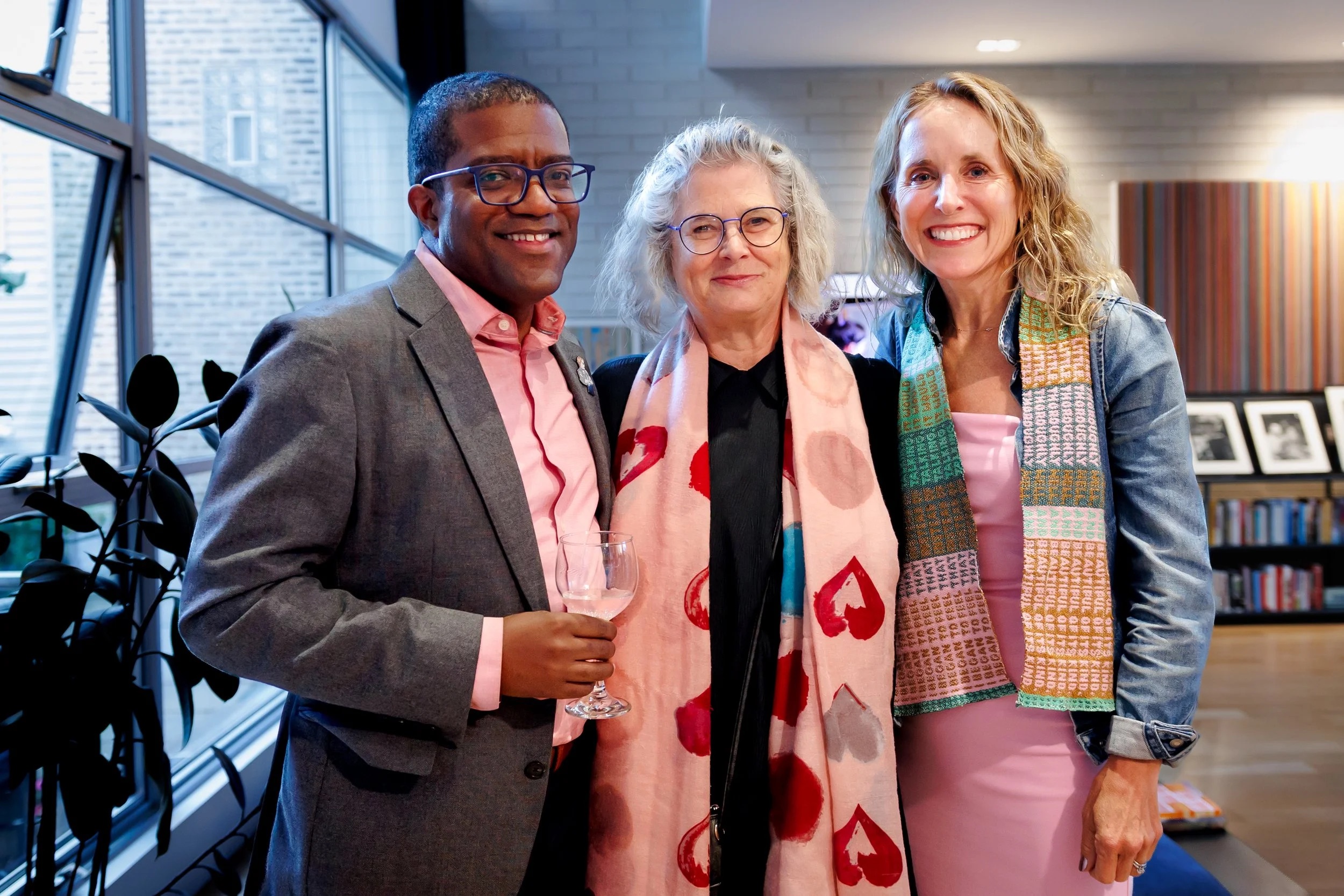 Three people stand together indoors, smiling at the camera. The man on the left holds a wine glass, and the two women beside him wear colorful scarves. Bookshelves and art are visible in the background.
