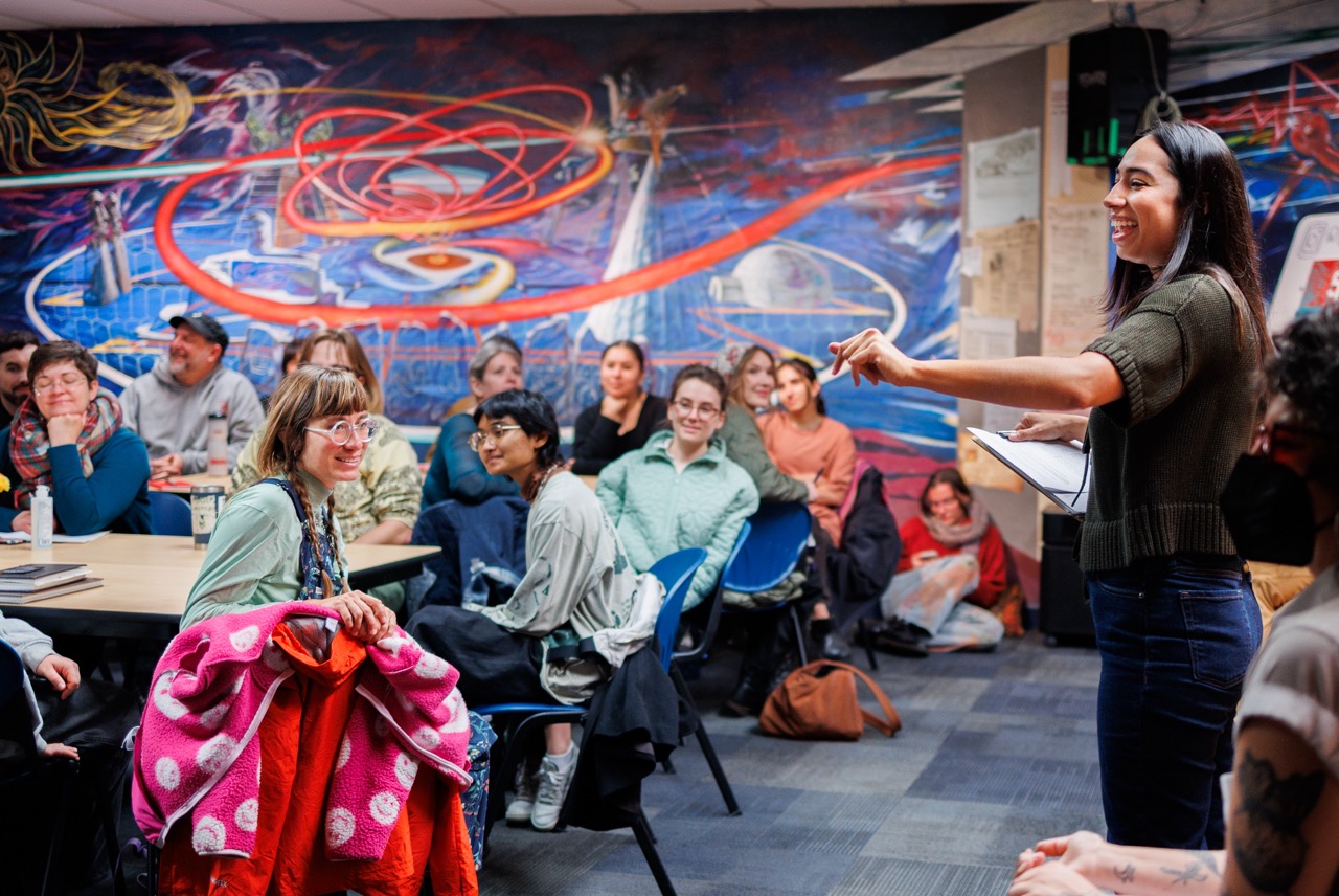 A woman stands and speaks to a smiling, attentive group seated in a colorful room with a vibrant mural on the wall behind them. Some people take notes while others listen closely.