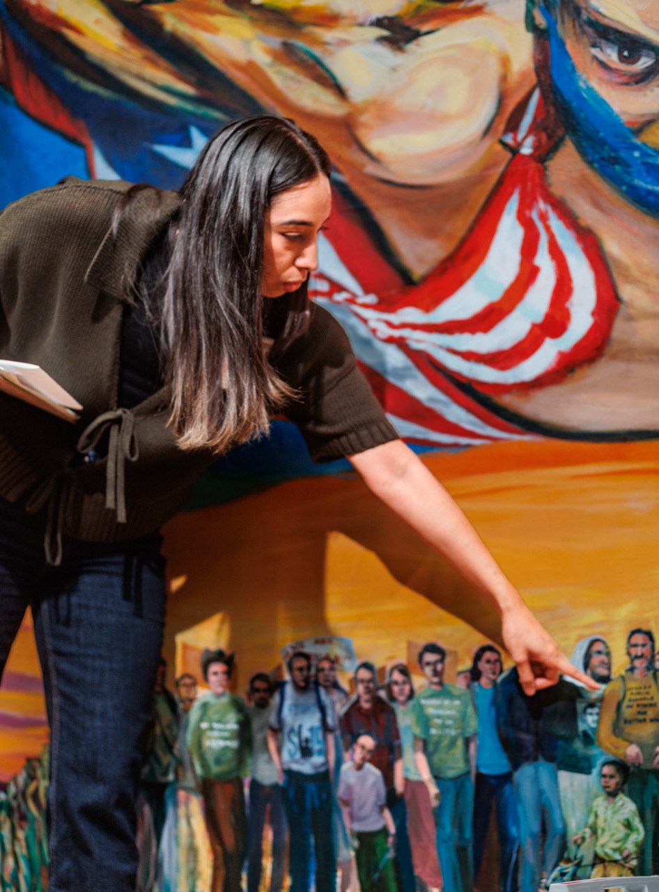 A woman with long dark hair points at a section of a colorful mural featuring a group of people standing together in front of bold, vibrant imagery. She holds a notebook in her other hand.